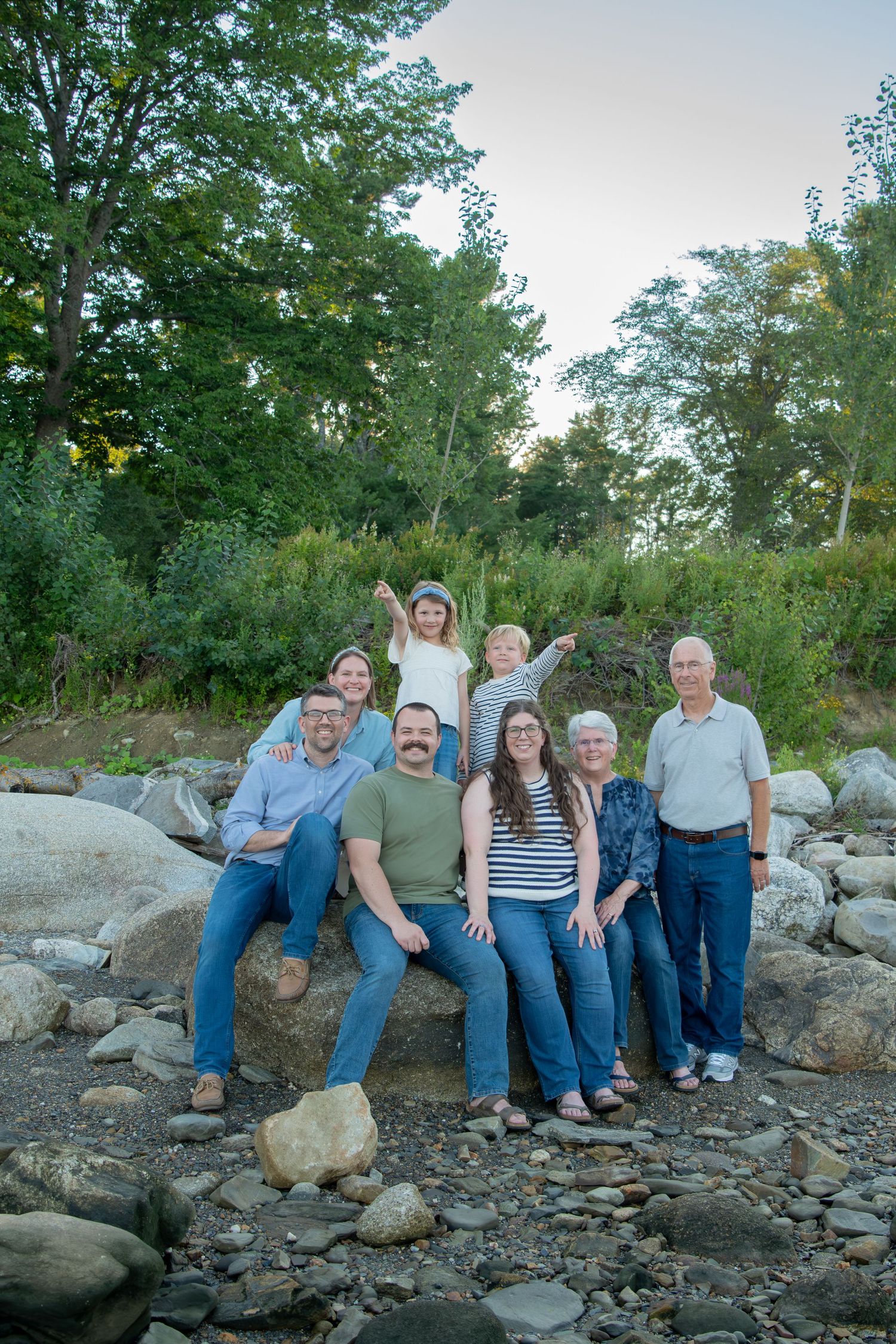 Multi-generational family of nine poses on rocky riverbank surrounded by lush green trees during golden hour outdoor portrait.