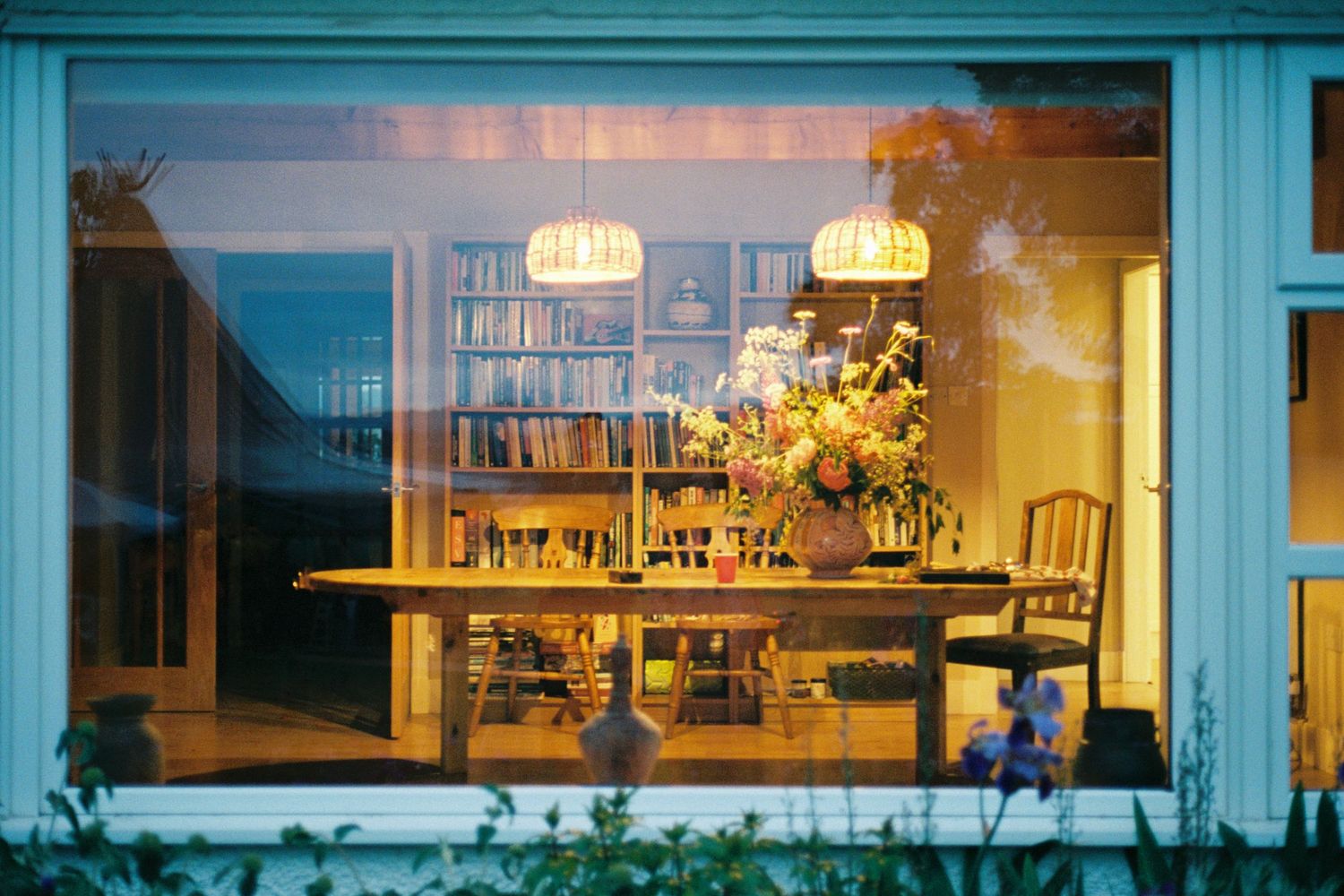 Cozy dining room viewed through large window at dusk, featuring wooden table, floral centerpiece, and bookshelf with warm lighting.