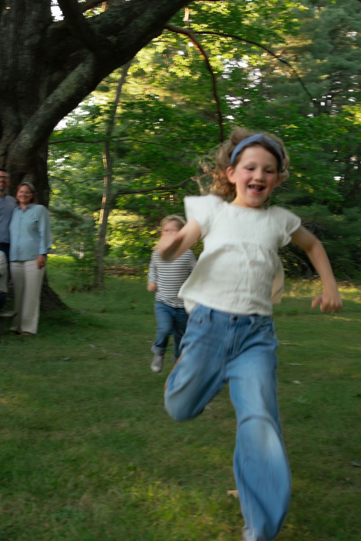 A joyful young girl with a headband runs across a green lawn, laughing, with children and adults visible behind her.