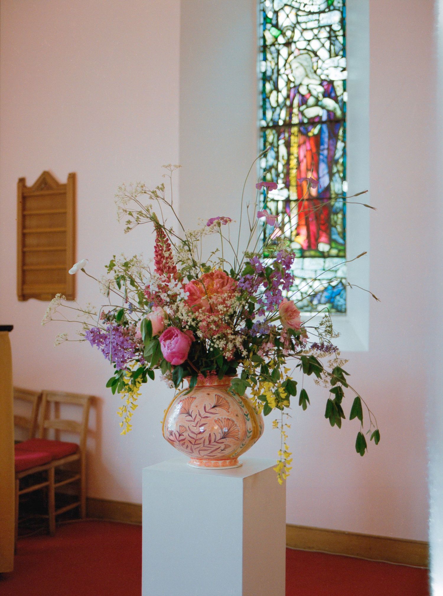 Colorful floral arrangement in a decorative vase on a white pedestal inside a church with stained glass window.