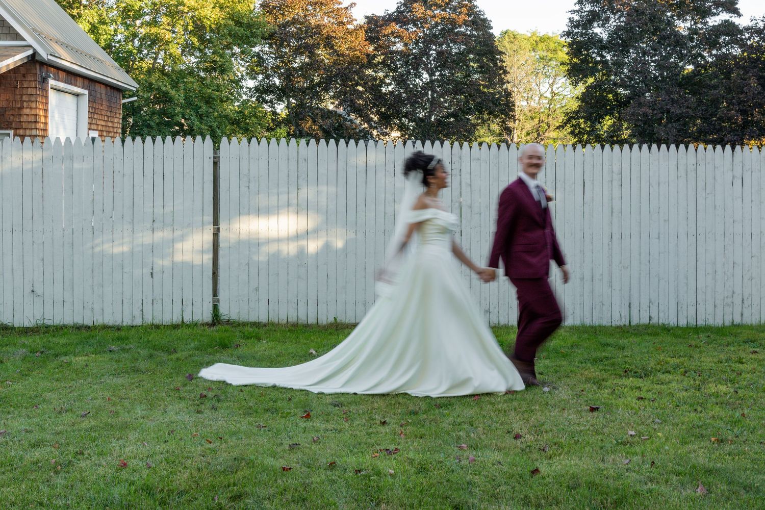 A bride in a white gown and groom in burgundy suit hold hands walking on green grass beside a white picket fence.