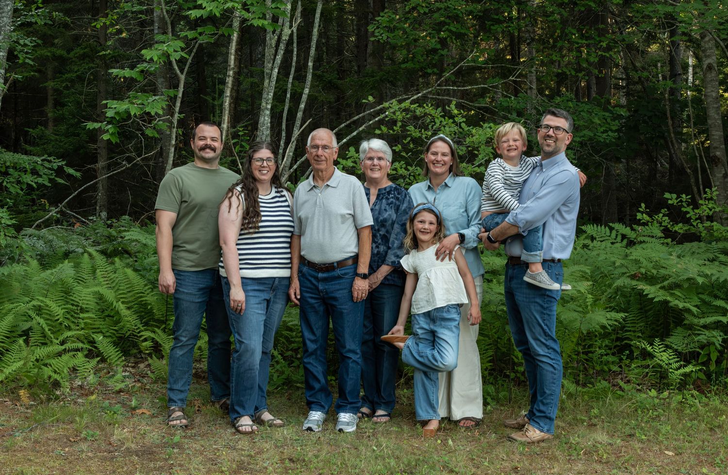 Multi-generational family group smiling together outdoors in a lush green forest setting for a portrait.