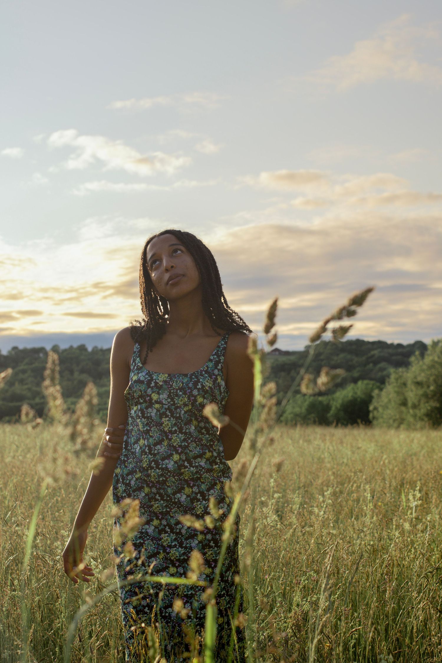 Woman in floral sundress standing in golden meadow at sunset, looking up dreamily with tall grass in foreground.