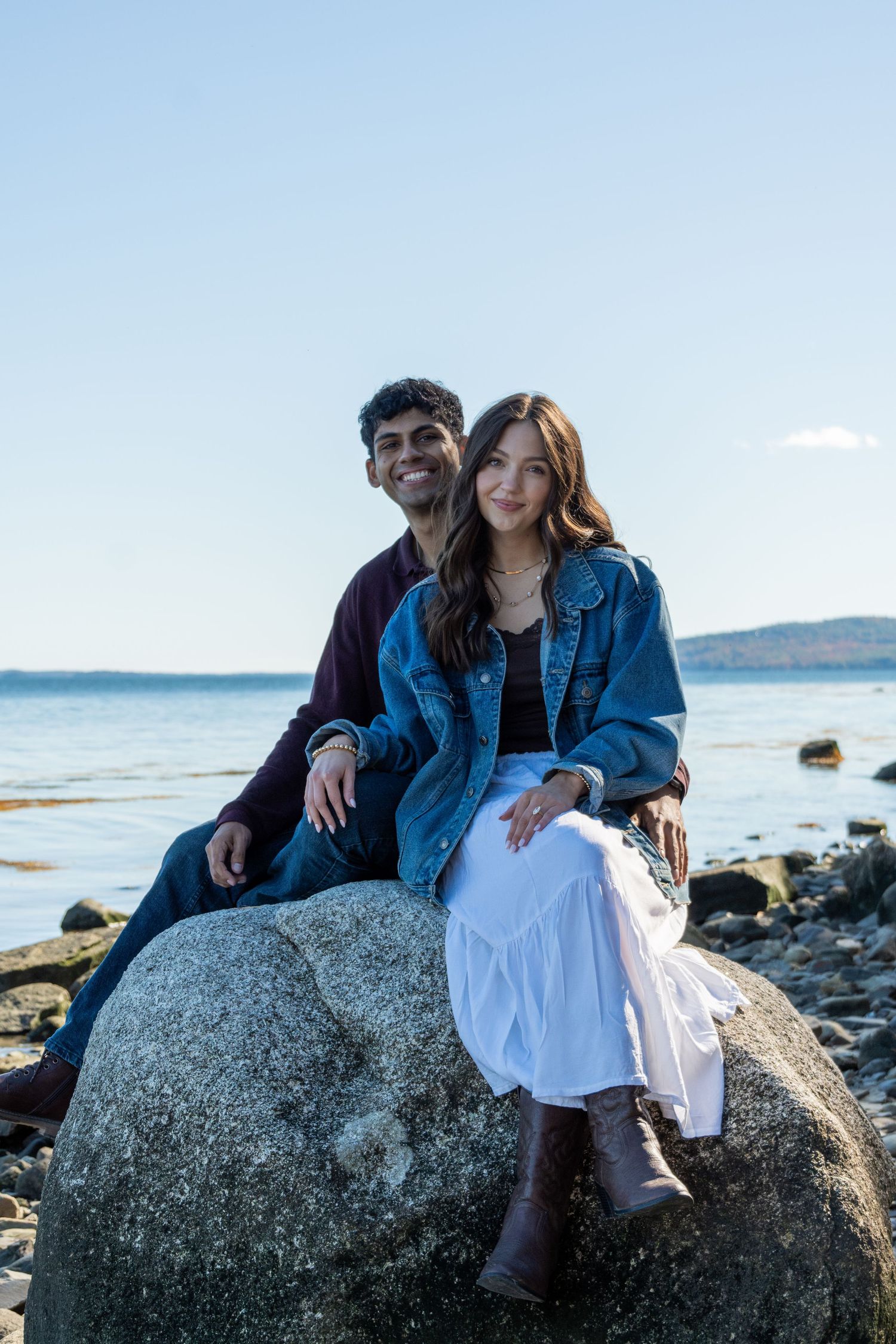 A couple sits together on a large rock by the ocean, smiling, wearing casual outfits on a sunny day.