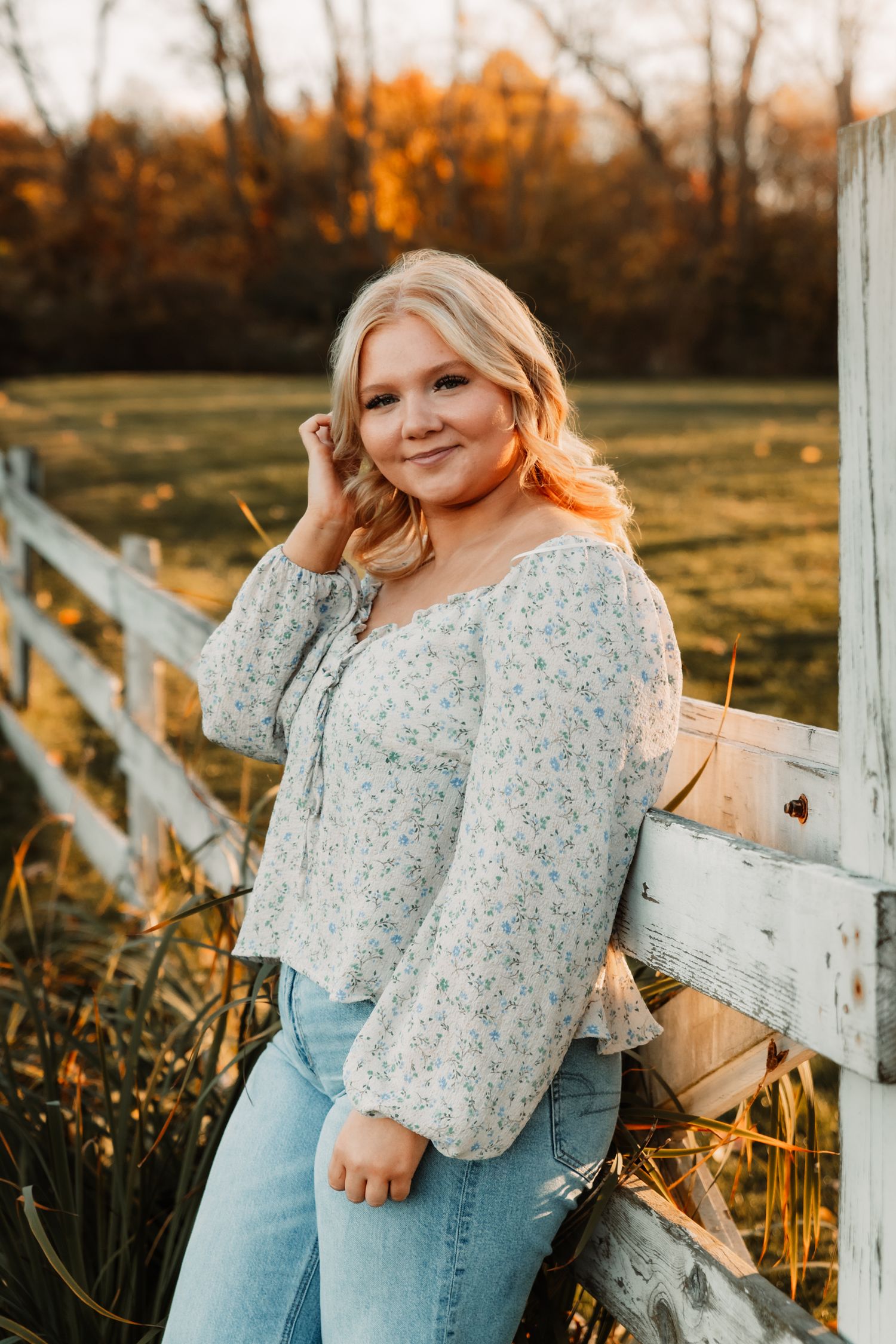Woman in floral blouse and jeans poses against rustic white fence during golden sunset in autumn field.