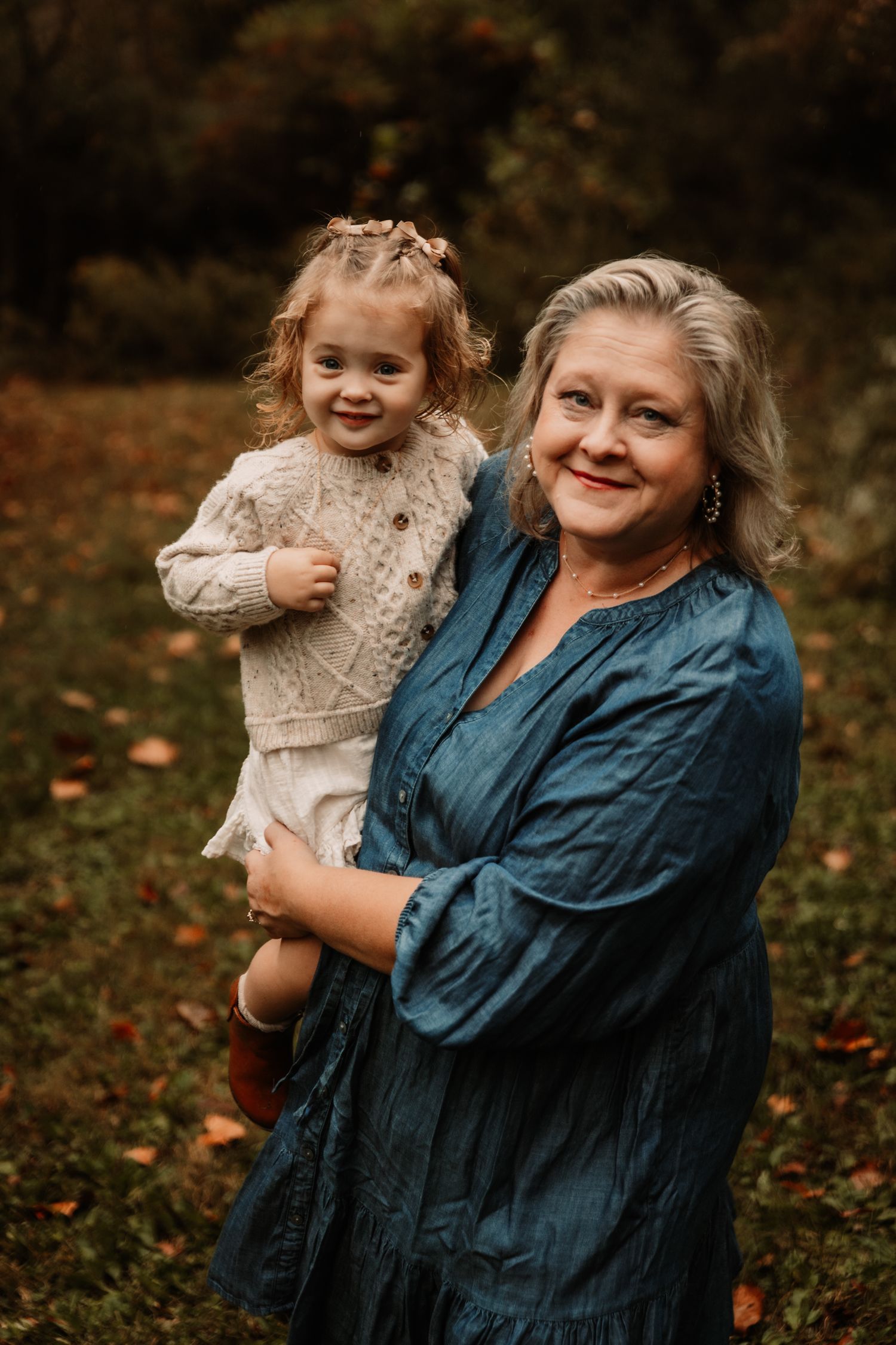 A child with blonde braids and a knit sweater smiles while being held in an autumn outdoor setting.