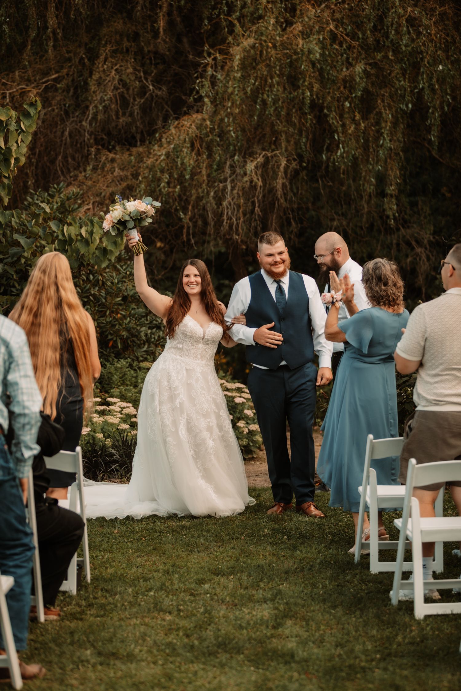 A joyful wedding ceremony outdoors with guests seated on white chairs while a couple celebrates walking down the aisle.