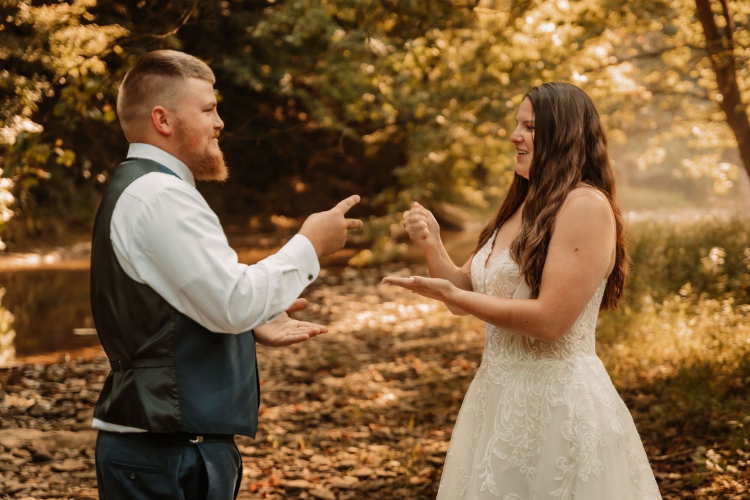 A couple in wedding attire laugh and point at each other playfully in a sunlit autumn forest setting.