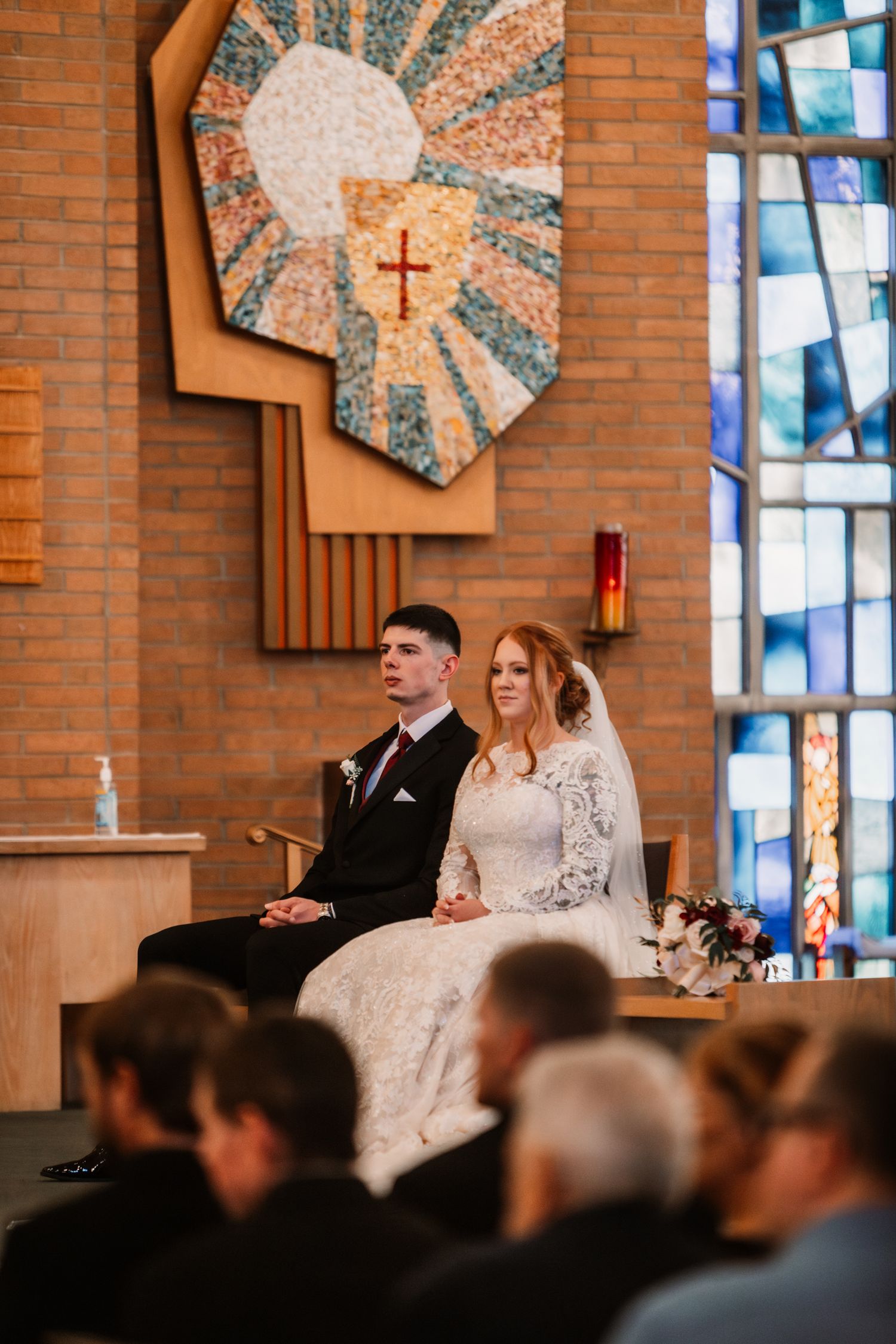 A traditional church wedding ceremony inside a brick chapel with stained glass windows and religious art on the wall.