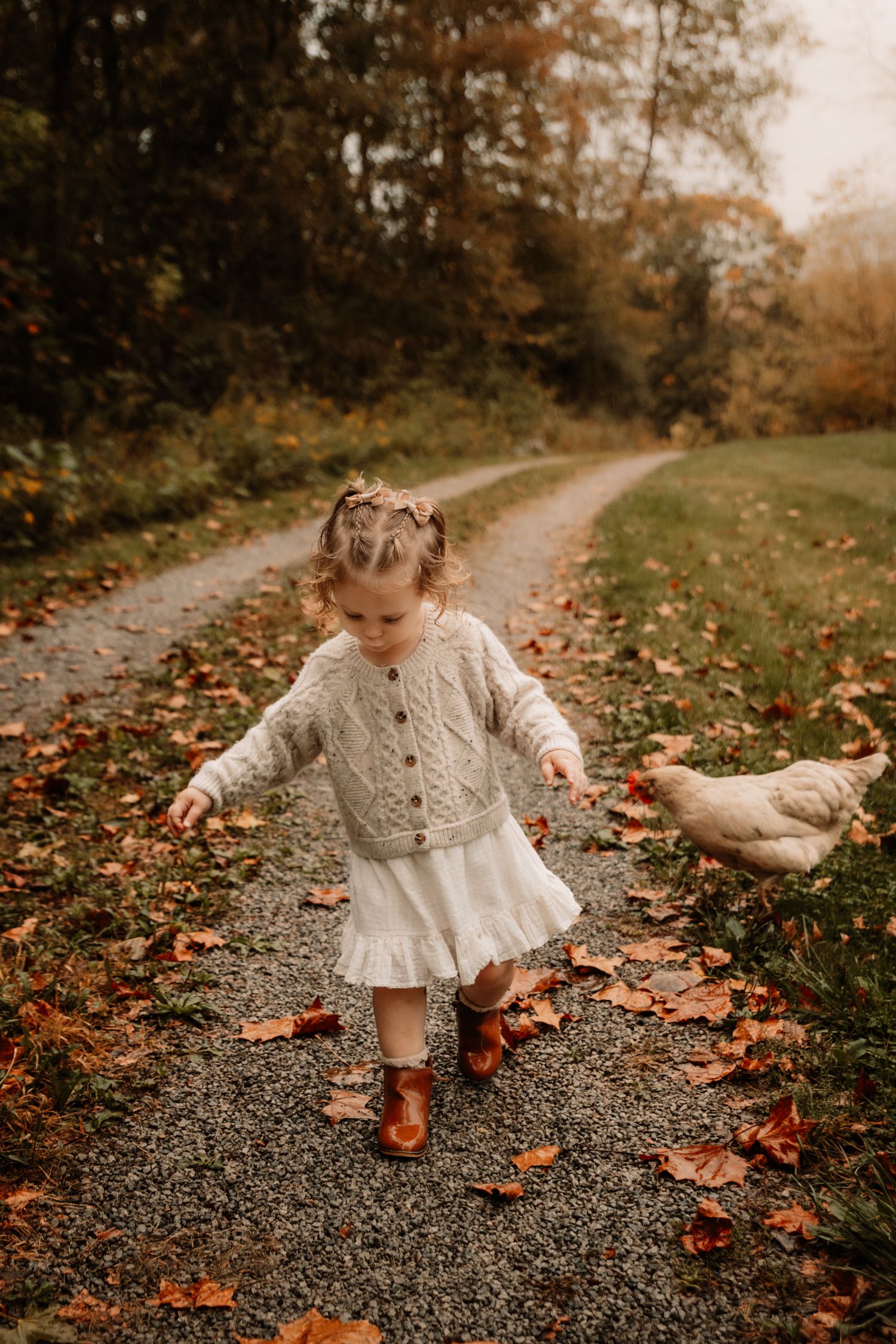 A toddler wearing a knitted sweater and white dress walks on a leaf-covered path during autumn while exploring nature.