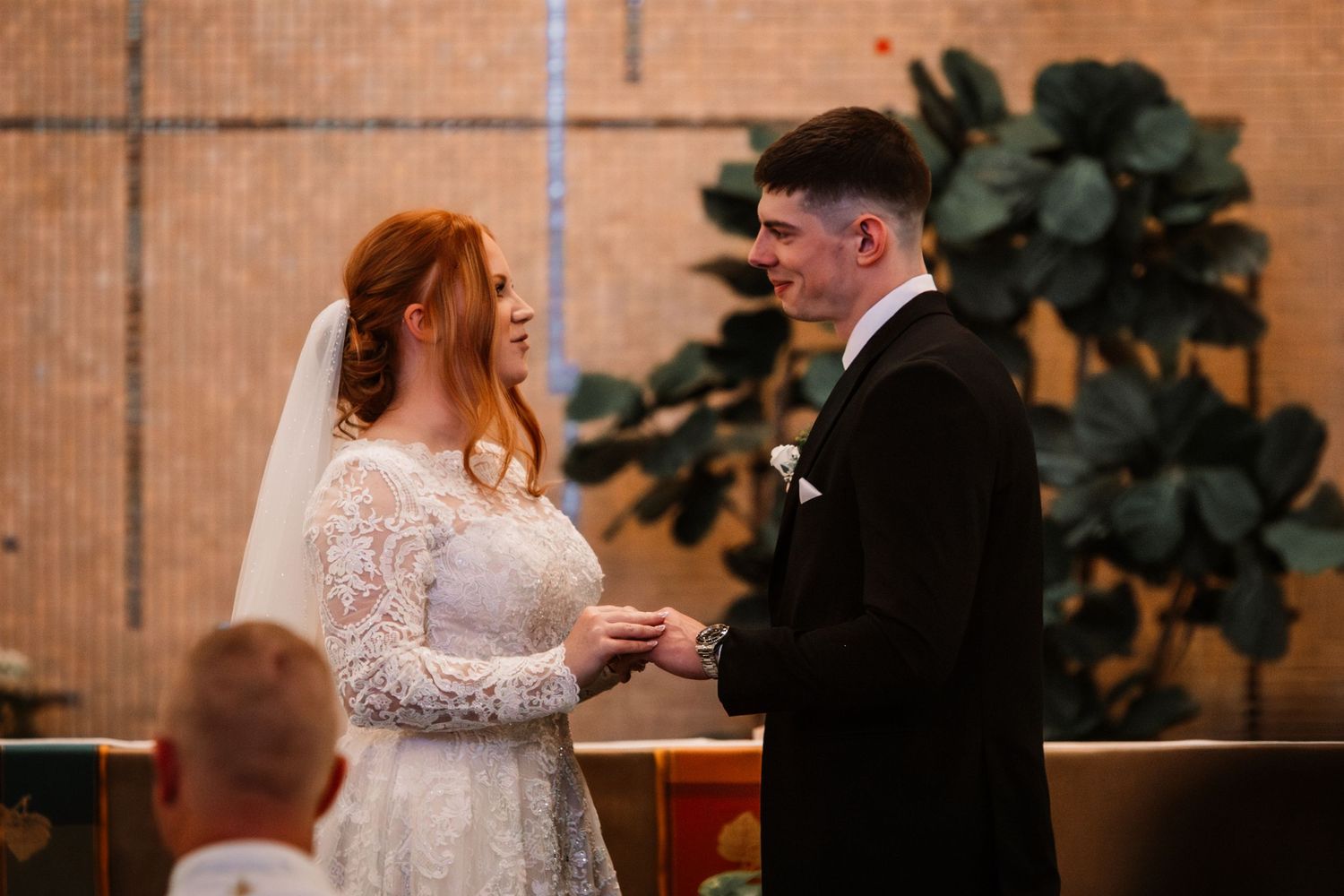 A couple exchanges rings during an intimate wedding ceremony in front of a brick wall with a large green plant.