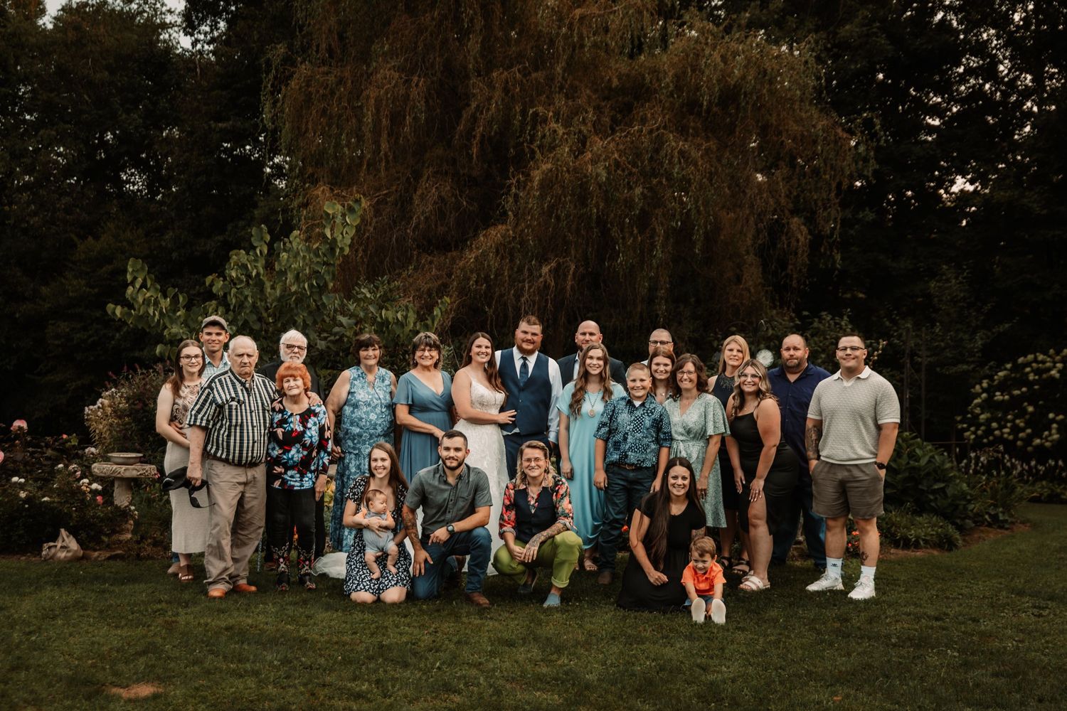 Large group of wedding guests in coordinated blue and casual attire pose together in a garden setting.