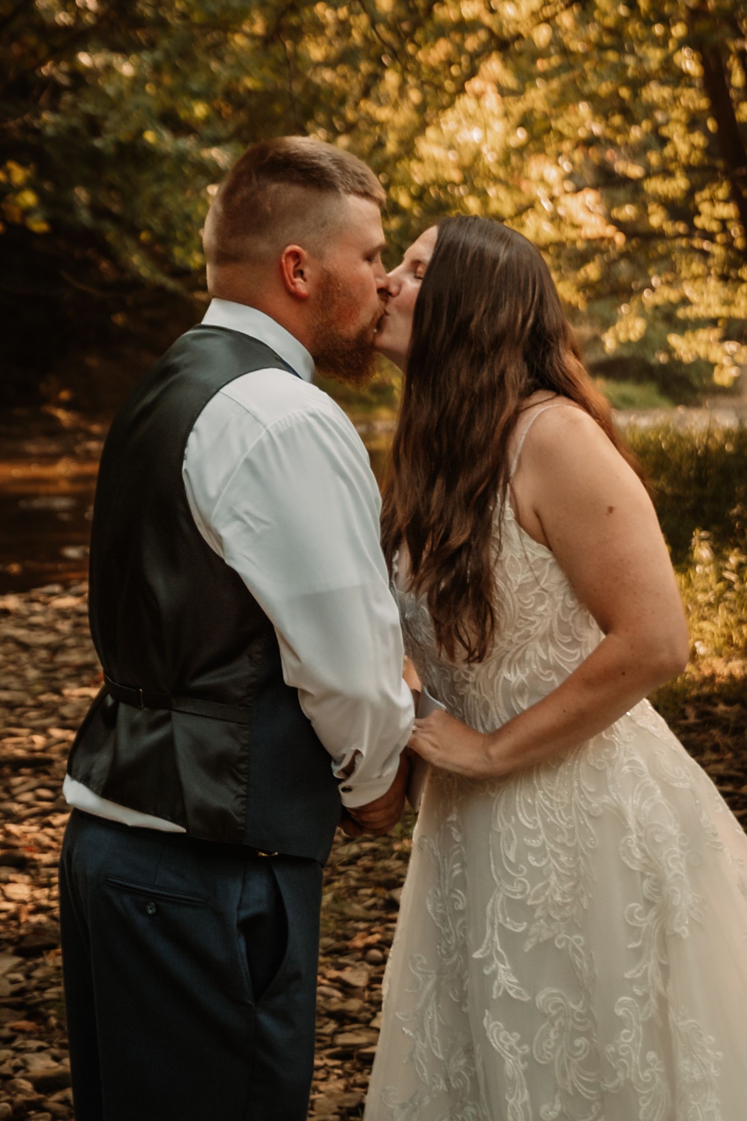 A romantic couple share a kiss in an autumn forest setting with golden sunlit trees in the background.