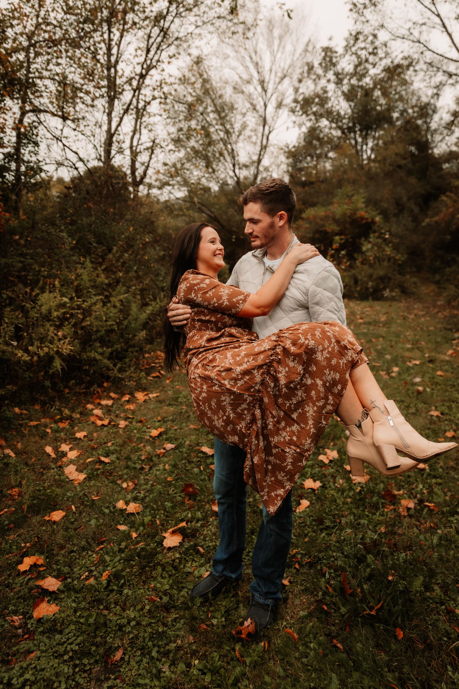 A romantic autumn photoshoot shows a couple embracing in a forest with fallen leaves as he carries her in his arms.
