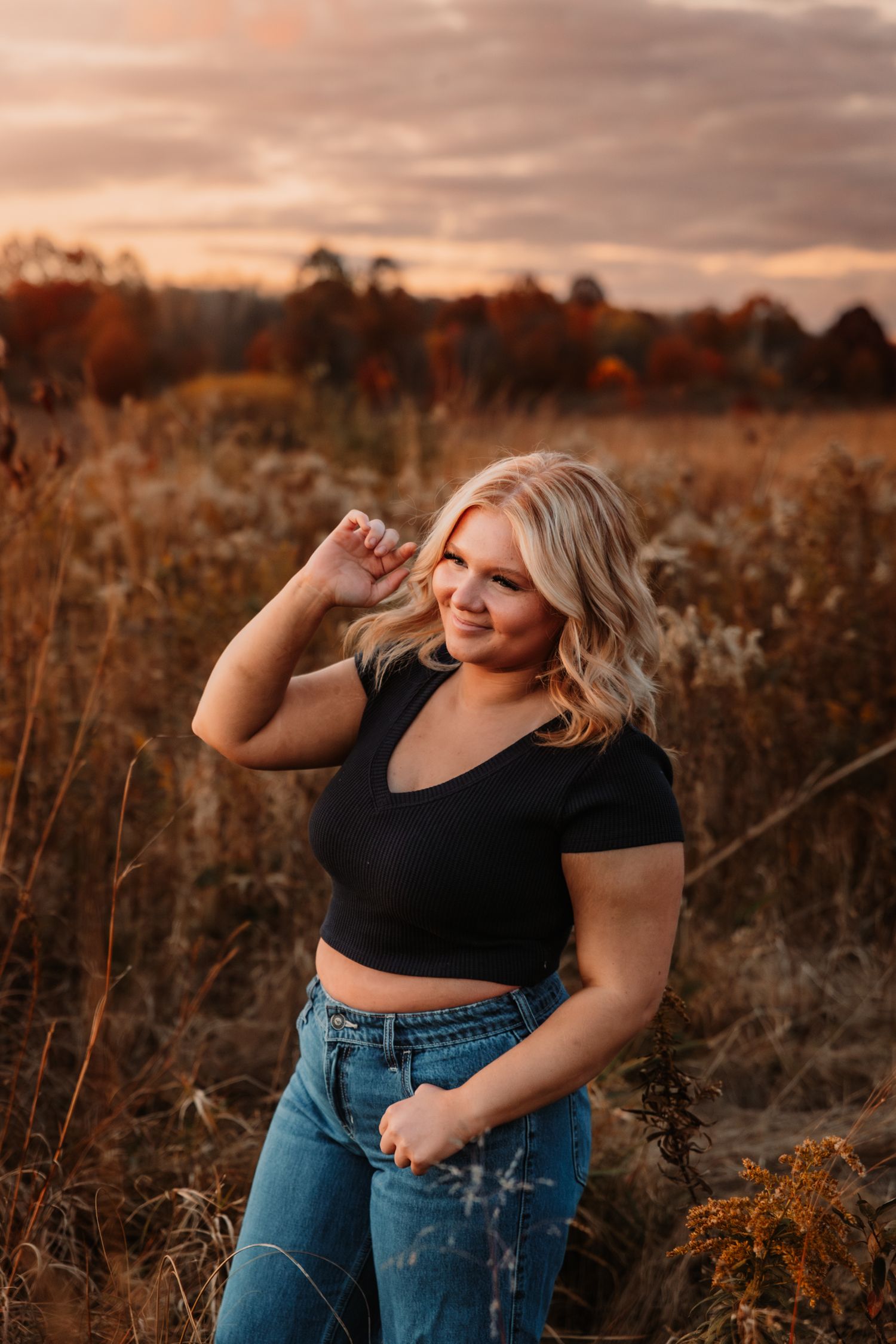 Golden sunset illuminates a rustic field with tall grass and autumn colors while a person poses in casual attire.