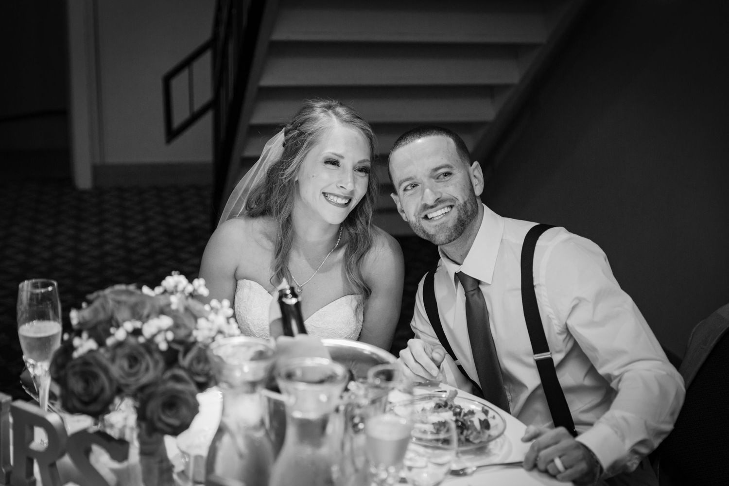 Black and white reception photo of newlyweds sharing joyful moment at decorated table.