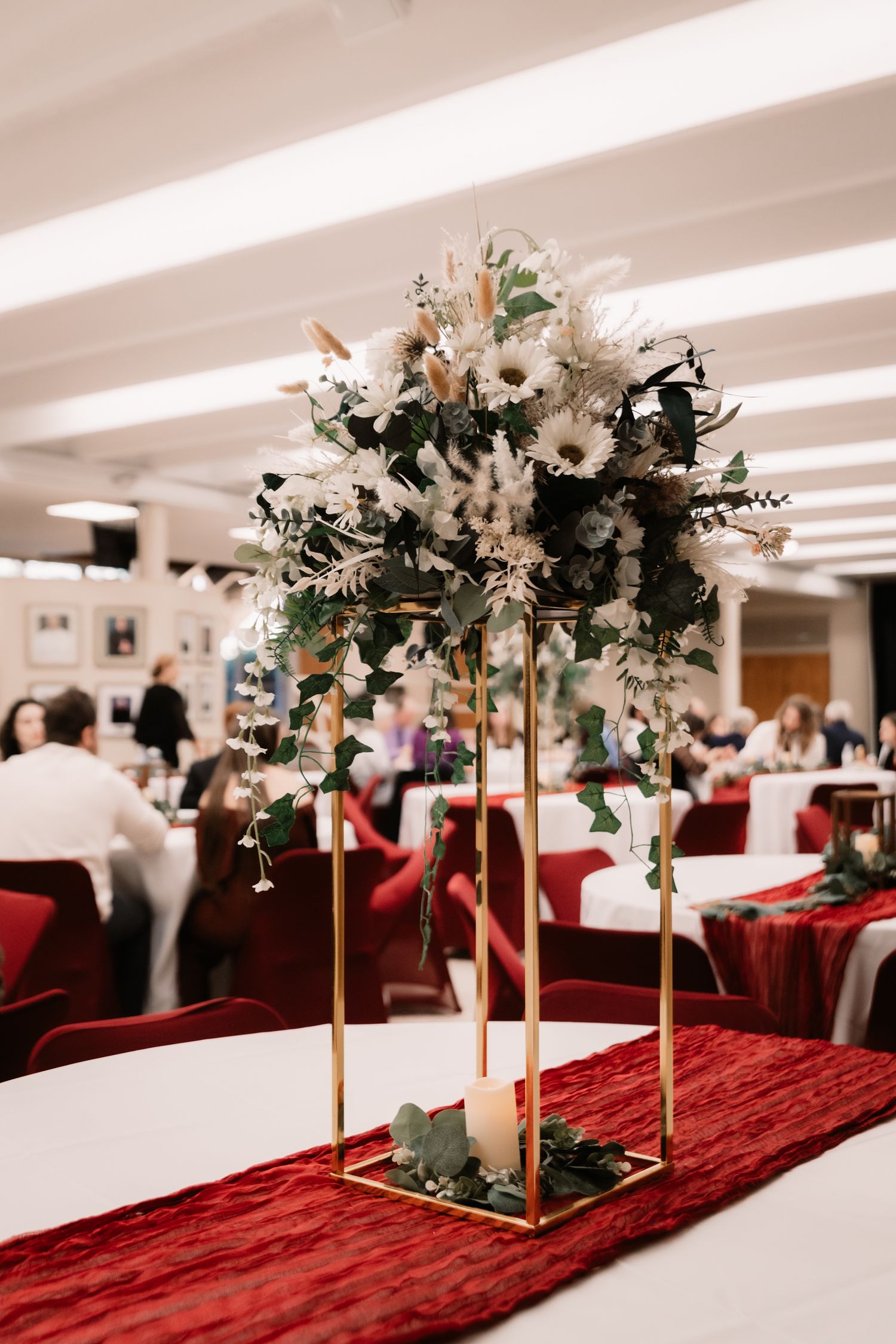 Elegant wedding centerpiece with white flowers and greenery on gold stands atop a red velvet table runner.