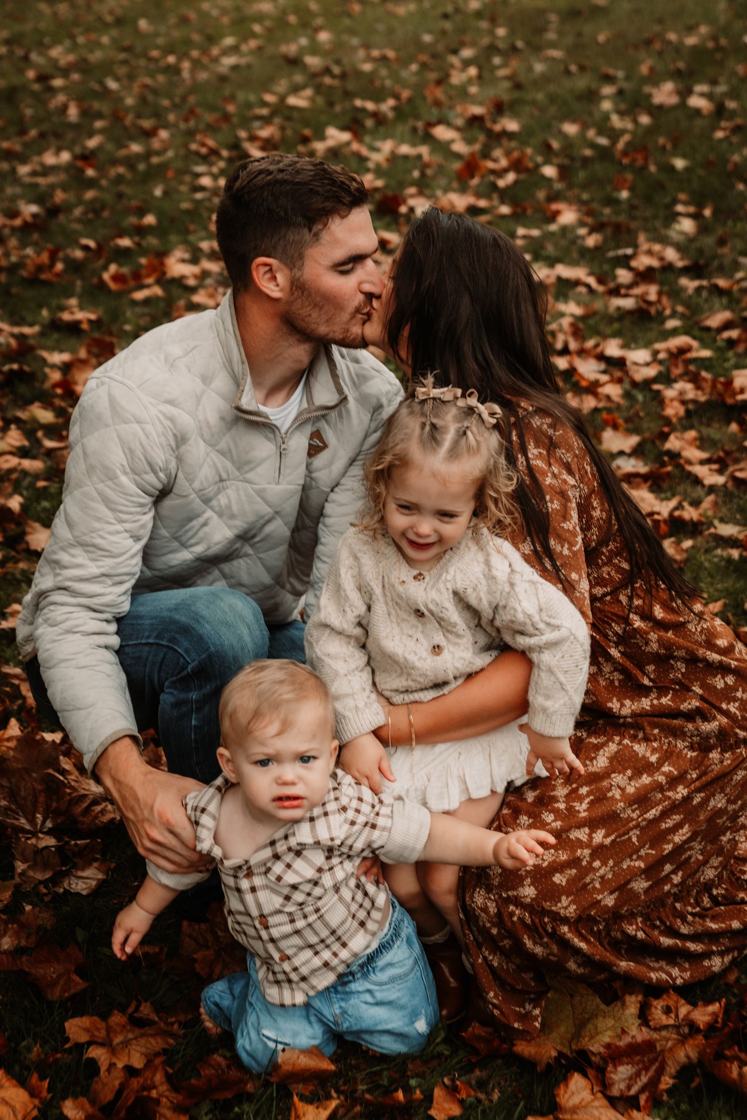 A family sits together on fallen autumn leaves wearing coordinating neutral outfits in warm earth tones.