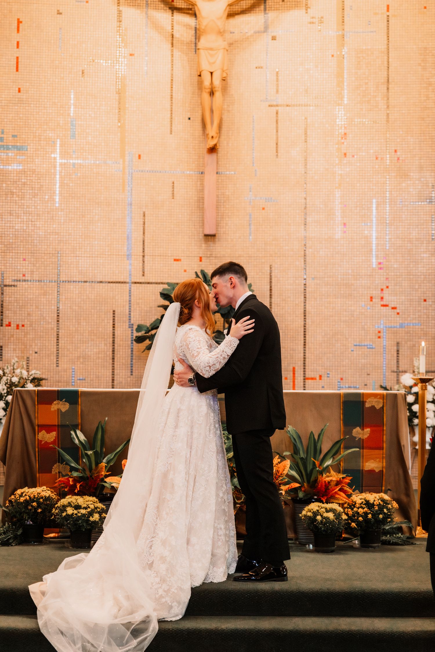 A couple shares an intimate moment at the altar during their wedding ceremony with autumn florals and a large crucifix.