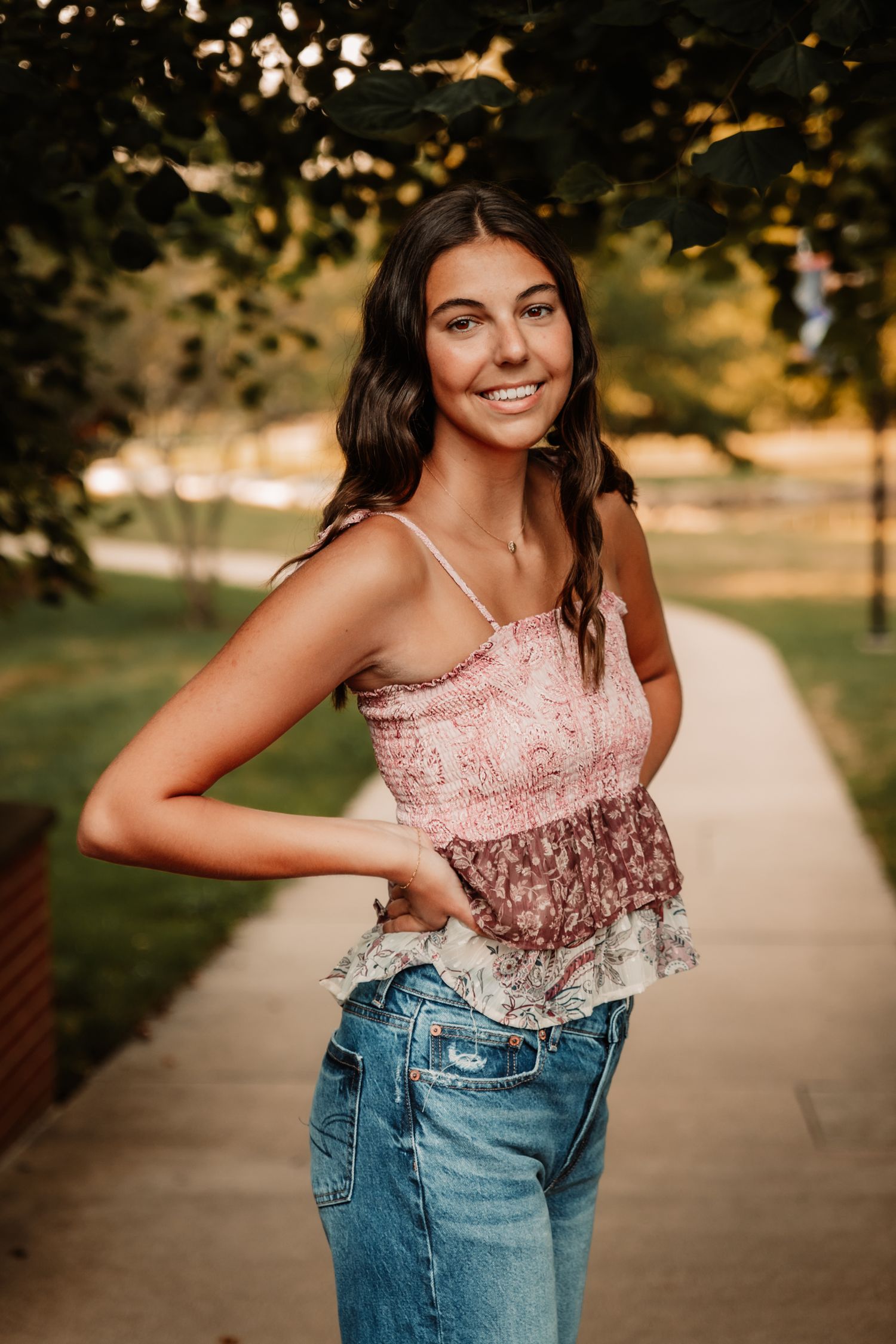 Someone wearing a pink lace camisole top and blue jeans poses on a sidewalk with trees and sunshine in the background.