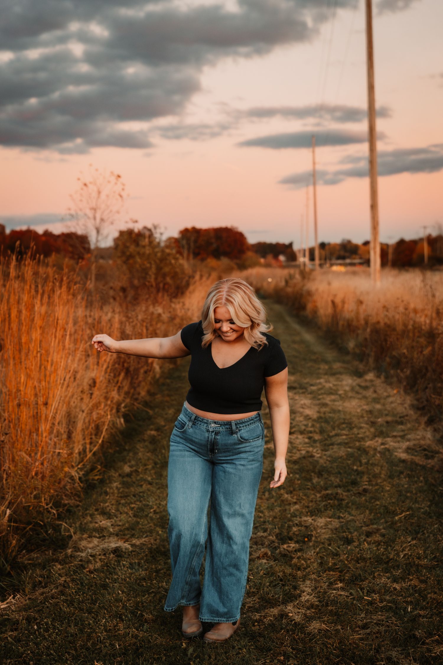 Person walking through a golden grassy field at sunset with dramatic cloudy skies and utility poles in background.