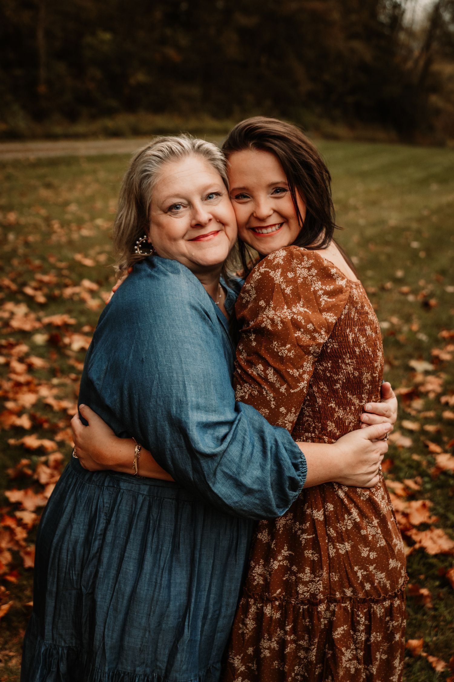 Two people wearing autumn dresses embrace and smile together on a fall day with leaves on the ground.