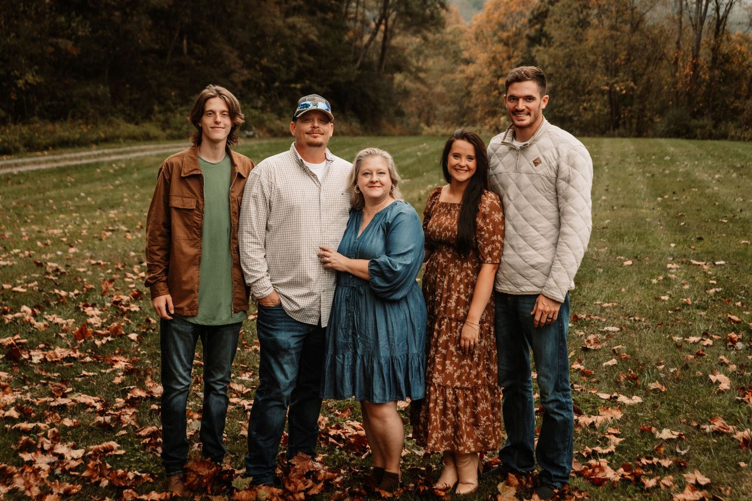 A group of five people pose together outdoors on fallen autumn leaves wearing coordinated casual outfits in earth tones.