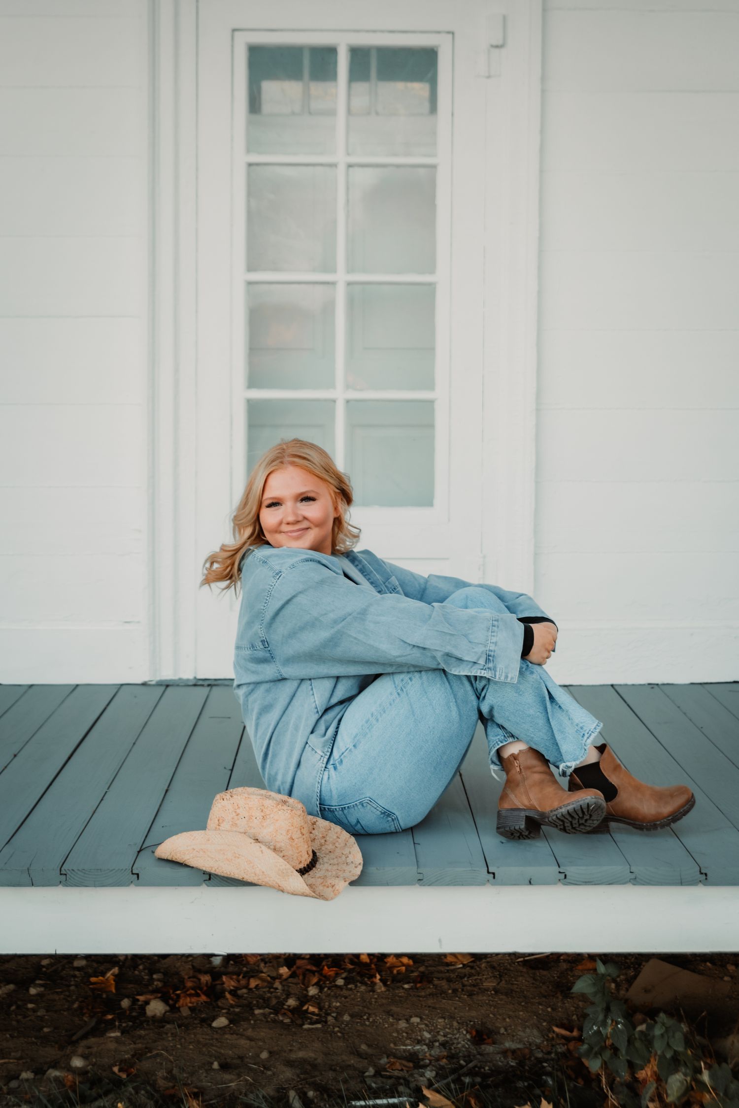 Casual portrait of someone in denim wearing brown boots sitting on white porch with cowboy hat beside them.
