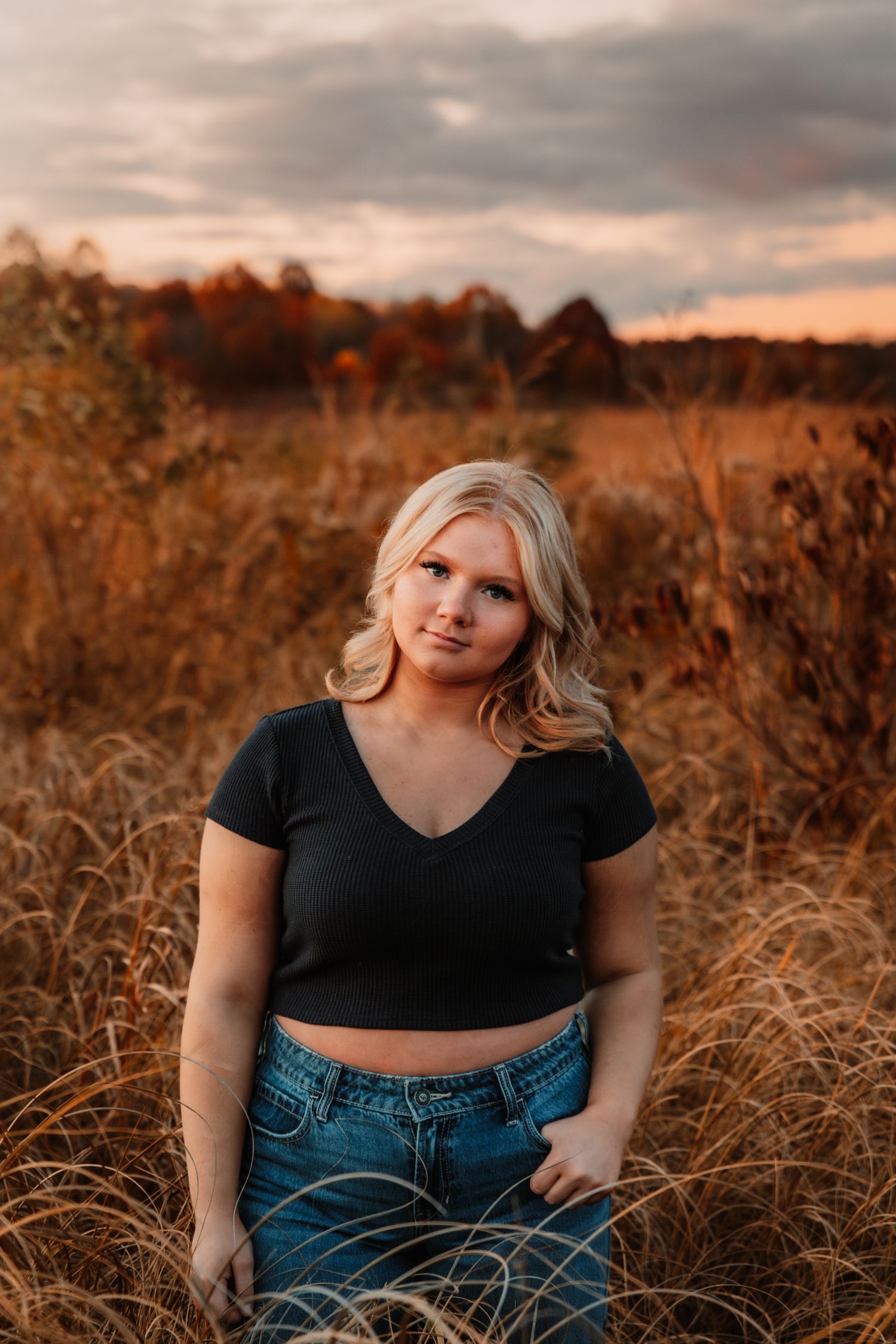 Outdoor portrait in golden autumn field with tall grass at sunset showing person in black crop top and jeans.