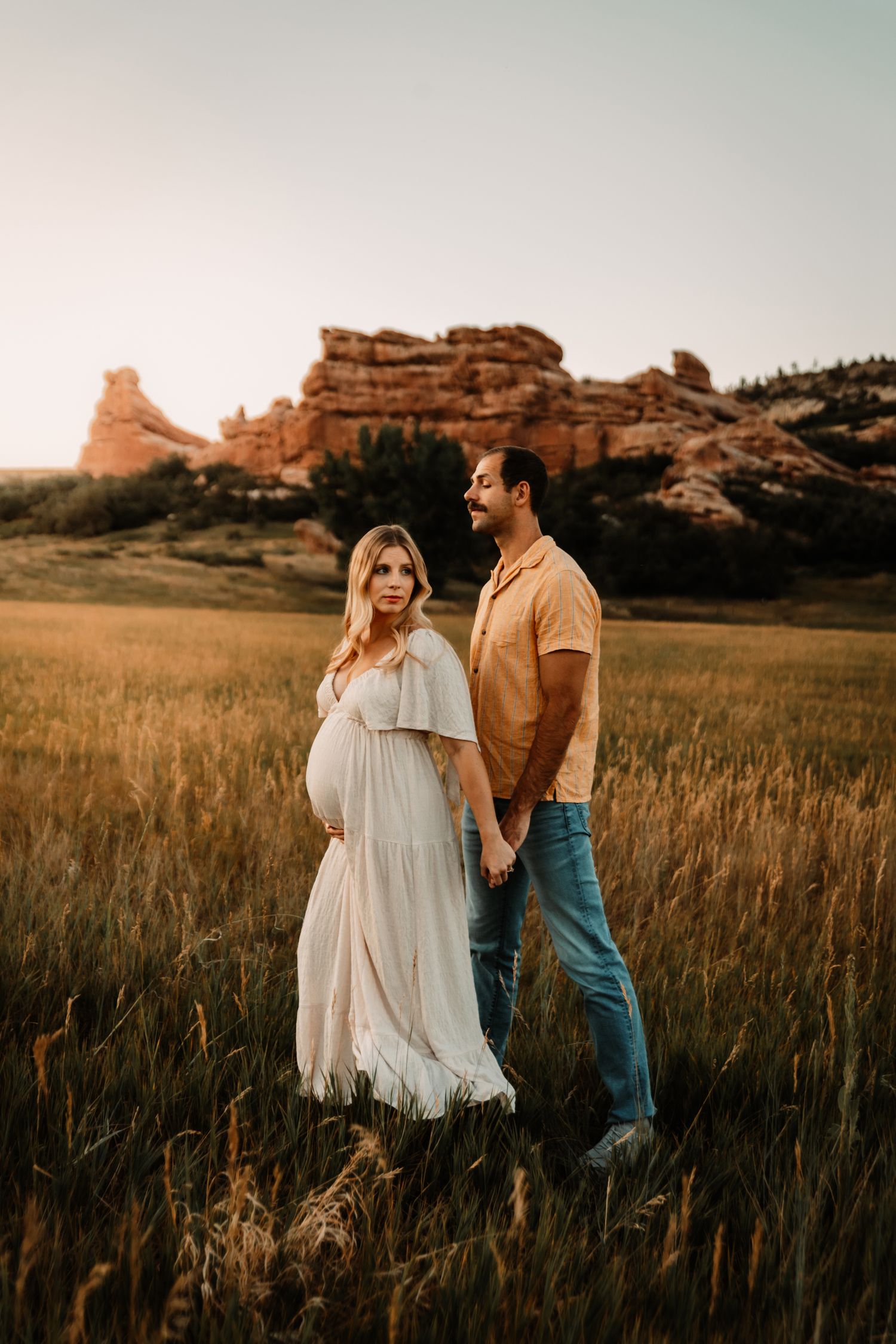 A couple stands together in a golden field at sunset with dramatic red rock formations in the background.