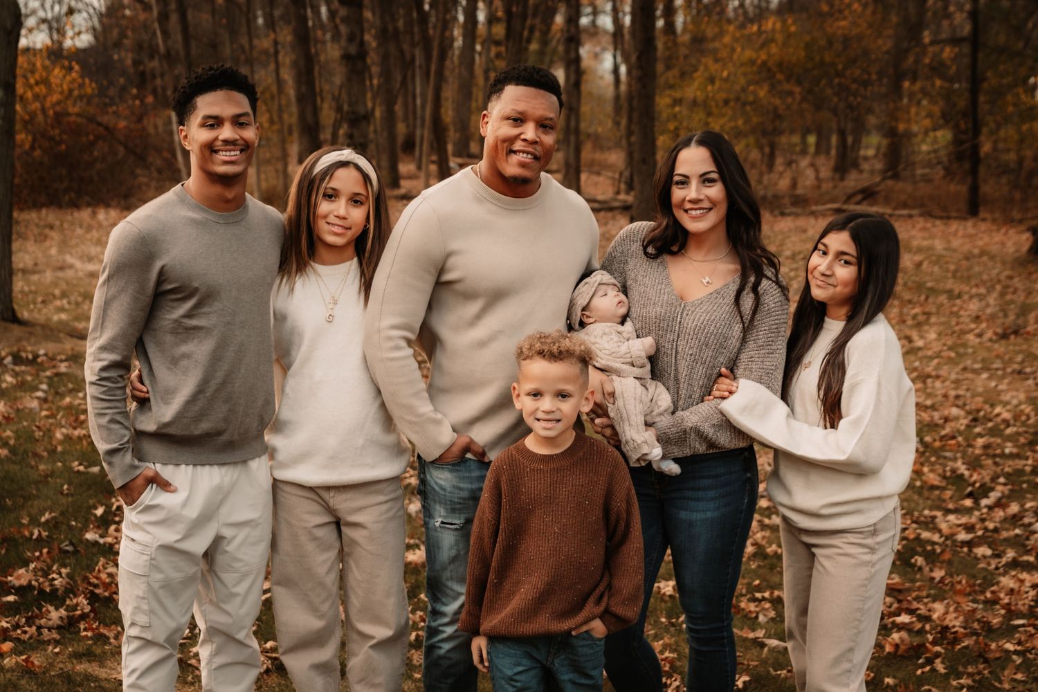 A group posed together outdoors during autumn with fallen leaves on the ground wearing casual sweaters in neutral tones.