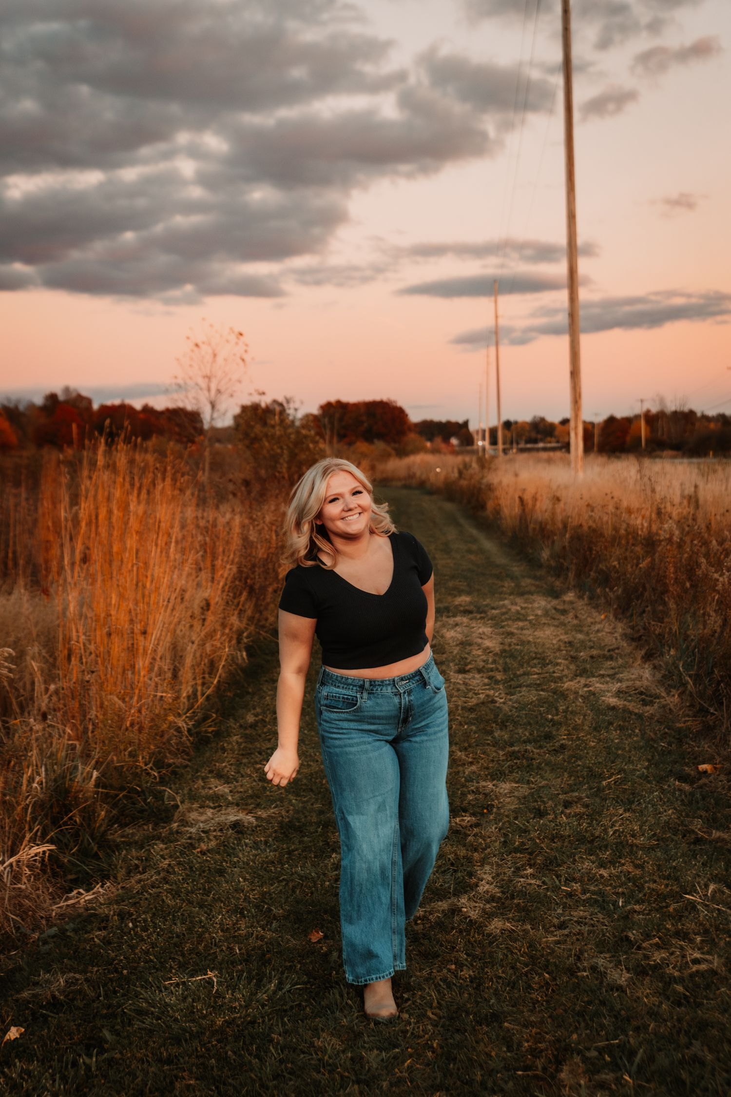 A person in a black crop top and jeans walks along a grassy path at sunset with golden light and dramatic clouds.