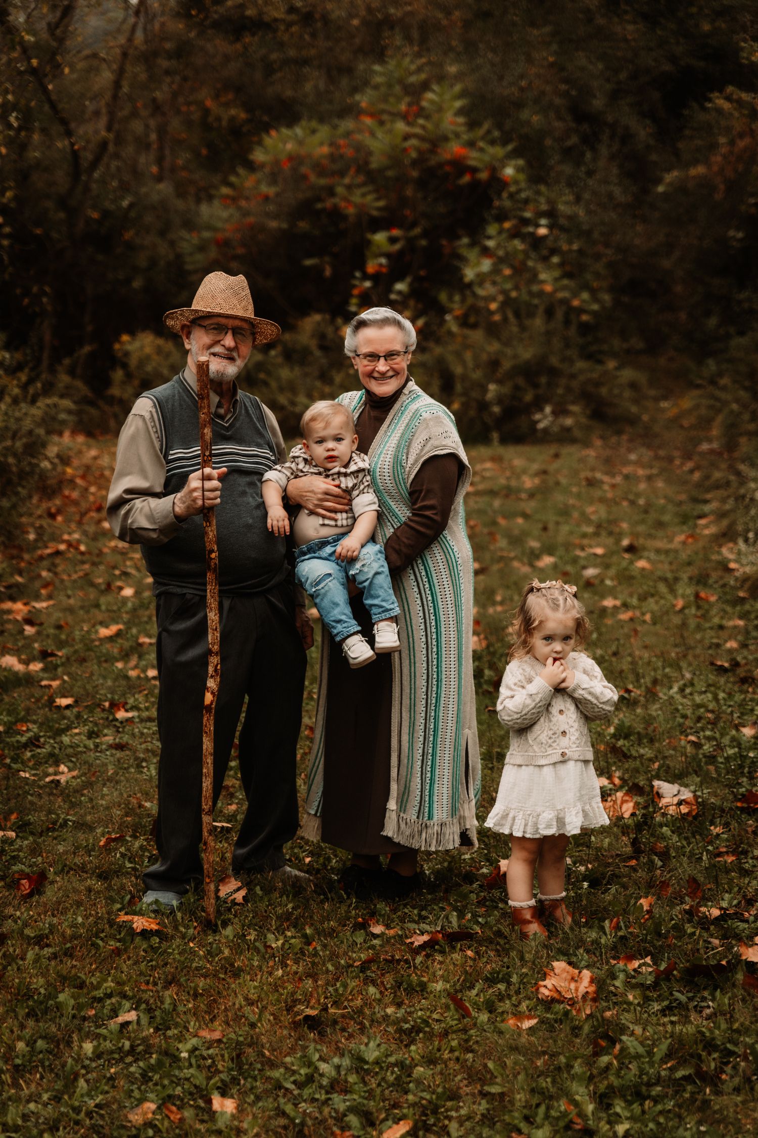 A family poses together outdoors in autumn, surrounded by fallen leaves and woodland scenery.