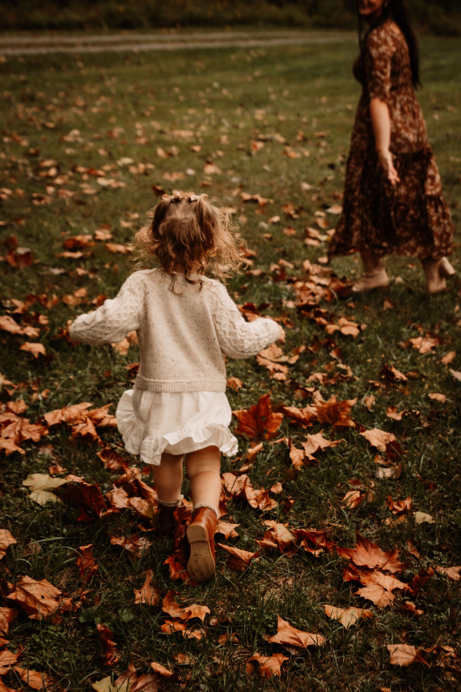 A child in a cream-colored dress and brown boots runs through autumn leaves scattered on grass.
