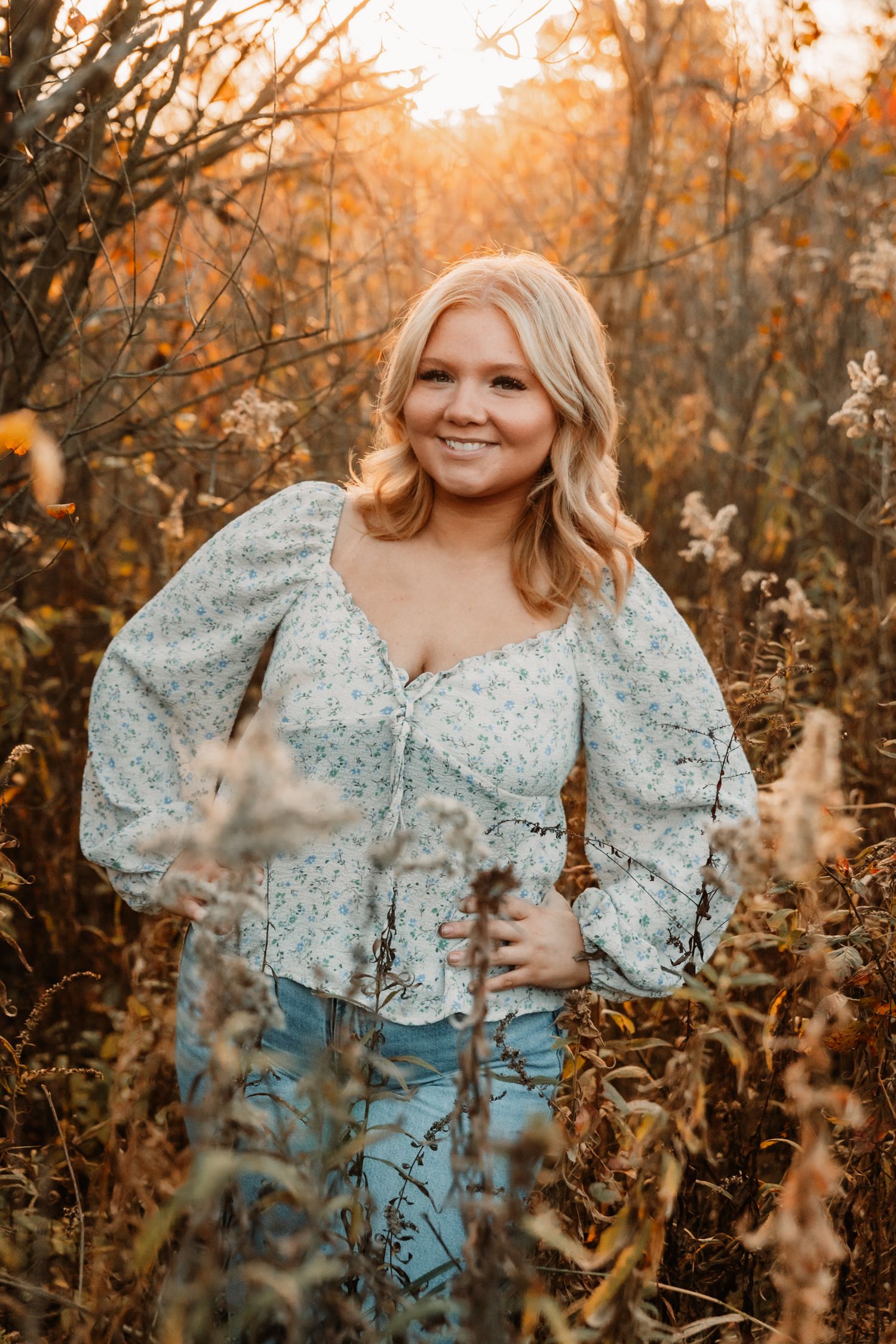 Woman in floral lace top poses outdoors in autumn field during golden hour sunset.