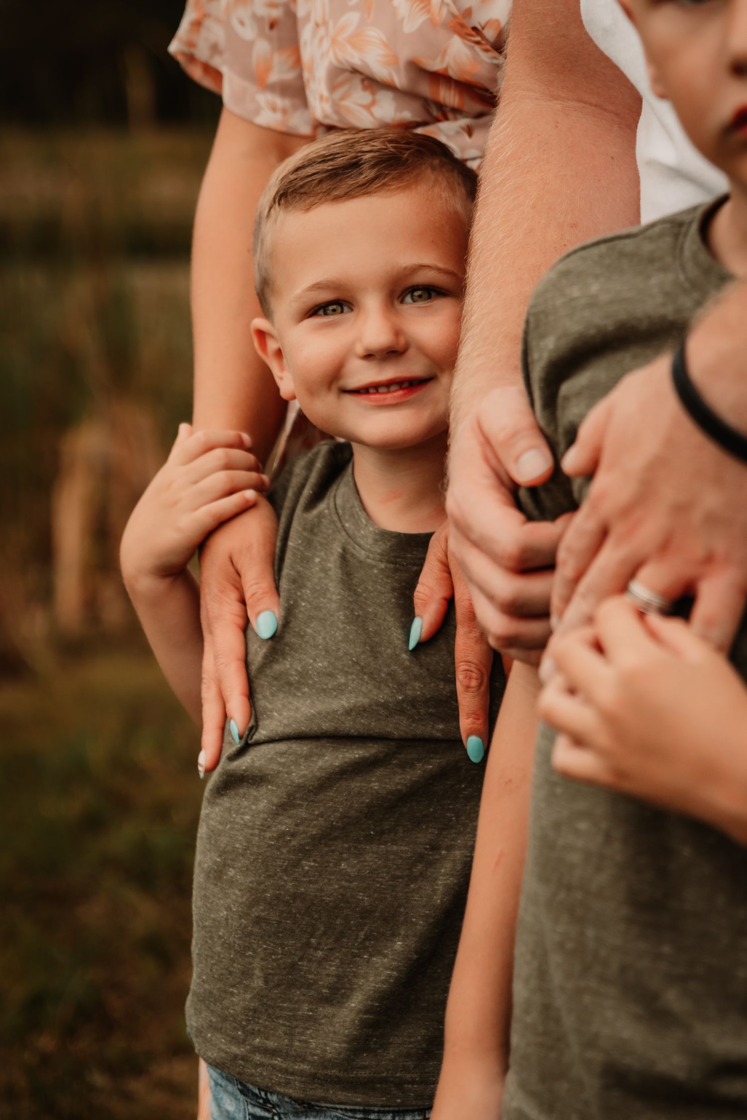 A young child in a grey tank top smiles while holding onto supportive hands around them in a warm outdoor setting.
