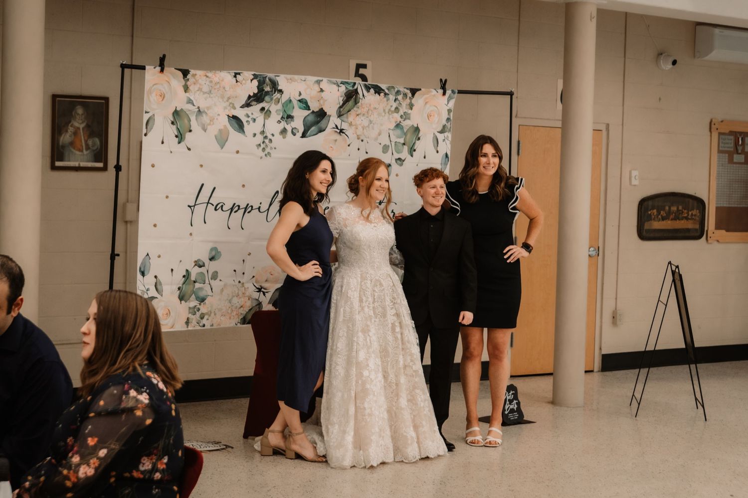 A group of people pose together at a reception in front of a floral backdrop with the word 'Happy' visible.