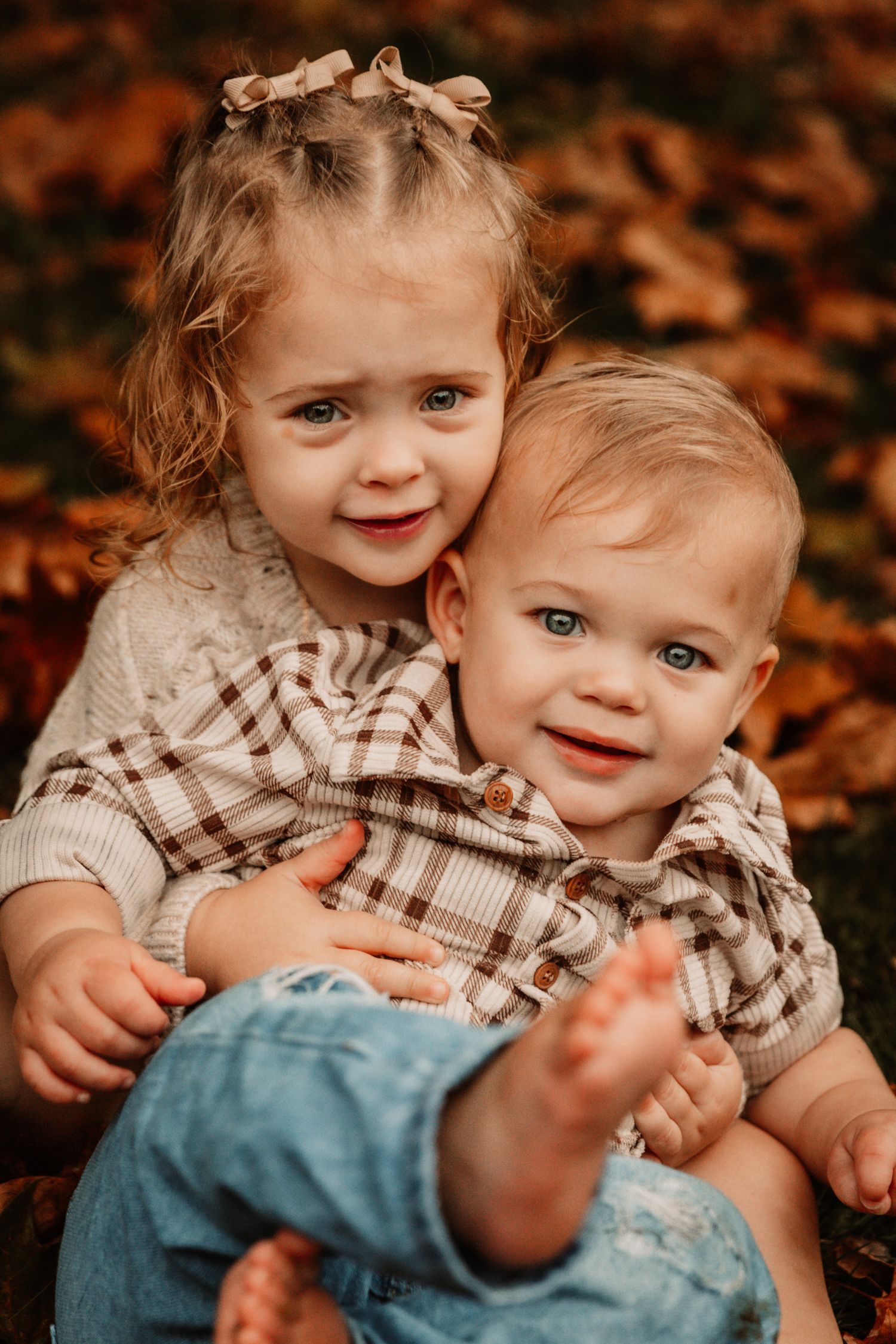 Two children in coordinated outfits sitting together among autumn leaves.