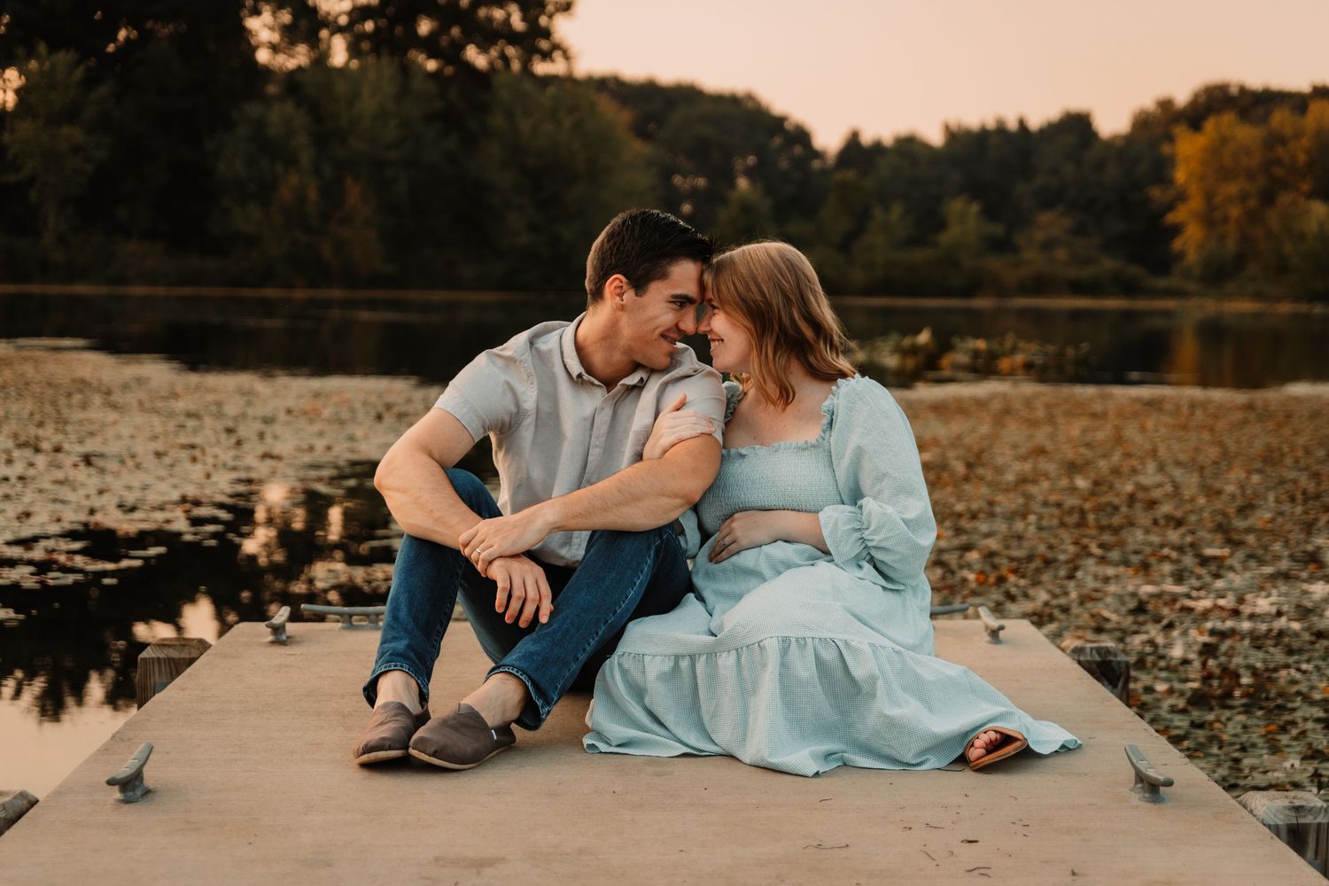 A couple sits together on a wooden dock by a tranquil lake during golden hour sunset.