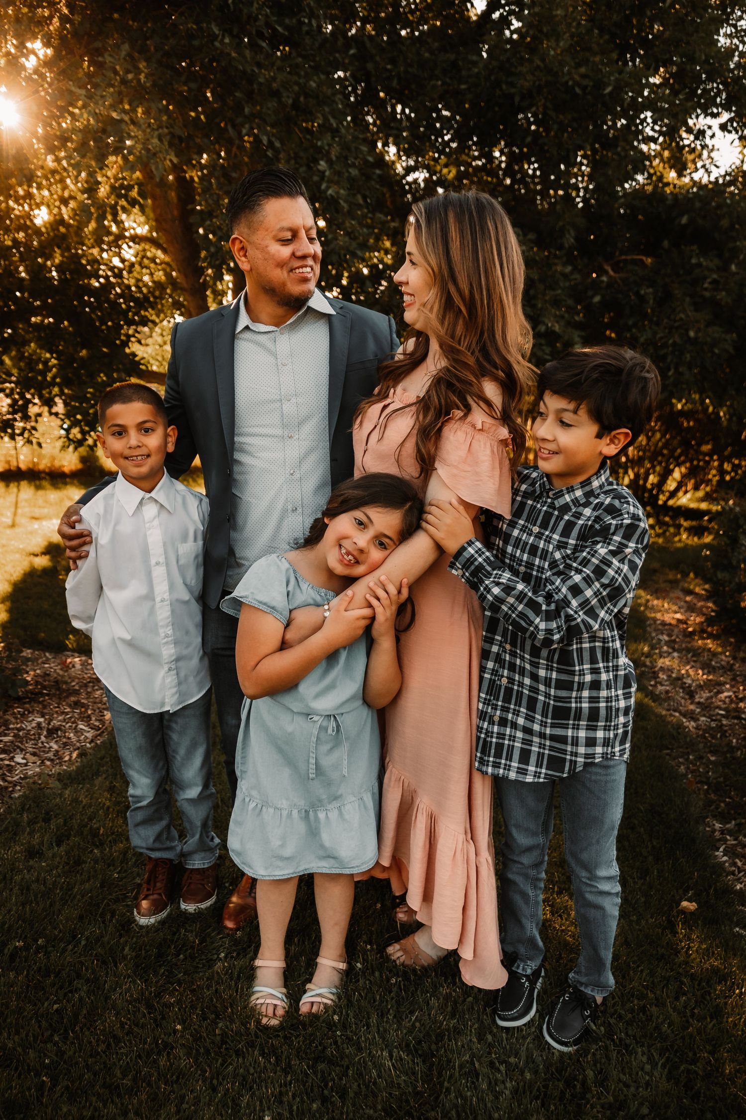 Family of five poses together outdoors at sunset with golden sunlight filtering through trees in the background.
