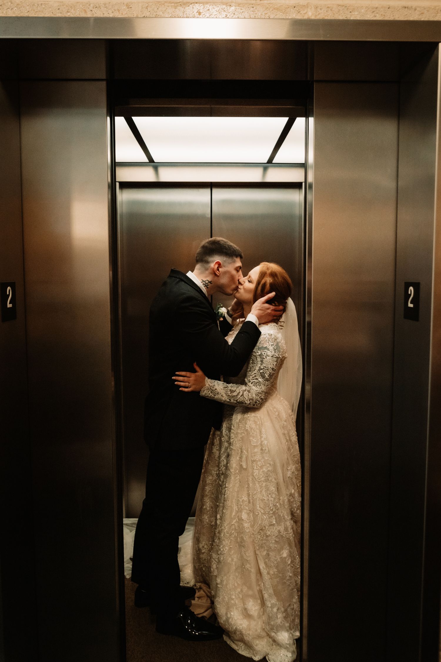 A romantic wedding moment captured in an elevator with a bride in a lace gown and groom in a black suit.