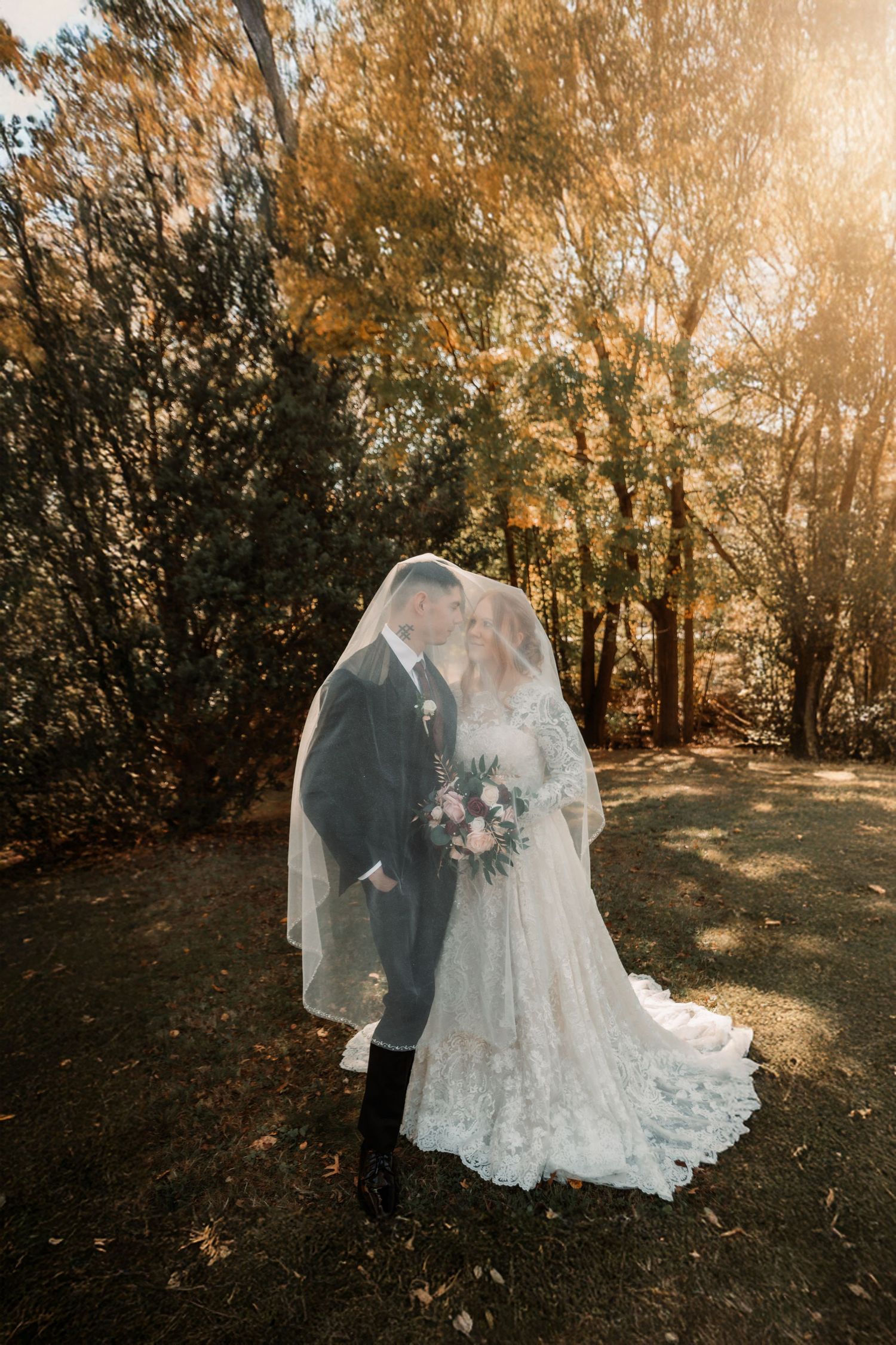 A couple in wedding attire shares an intimate moment in a sunlit autumn forest setting.