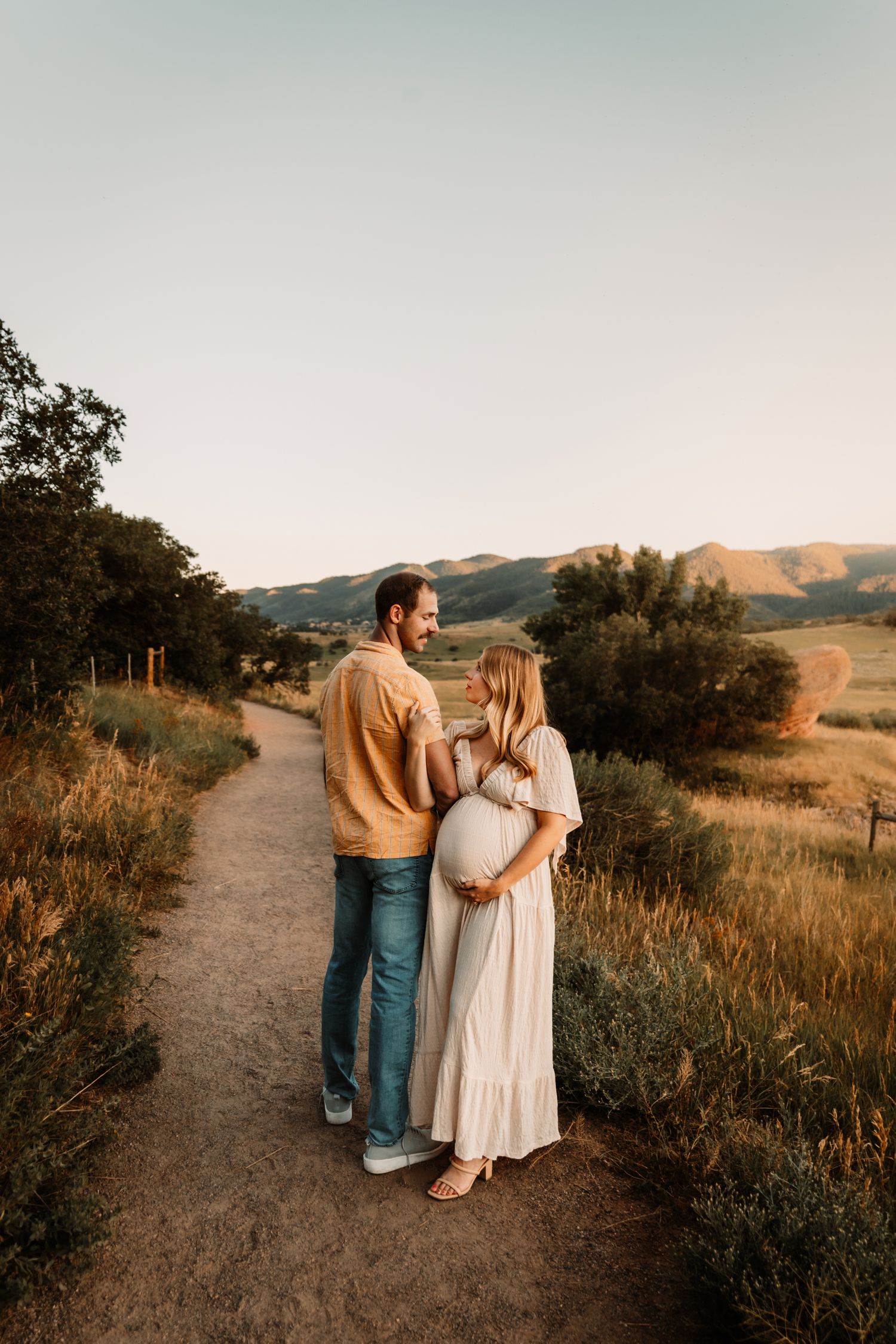 A couple stands together on a dirt path at sunset with golden hills and trees in the background.