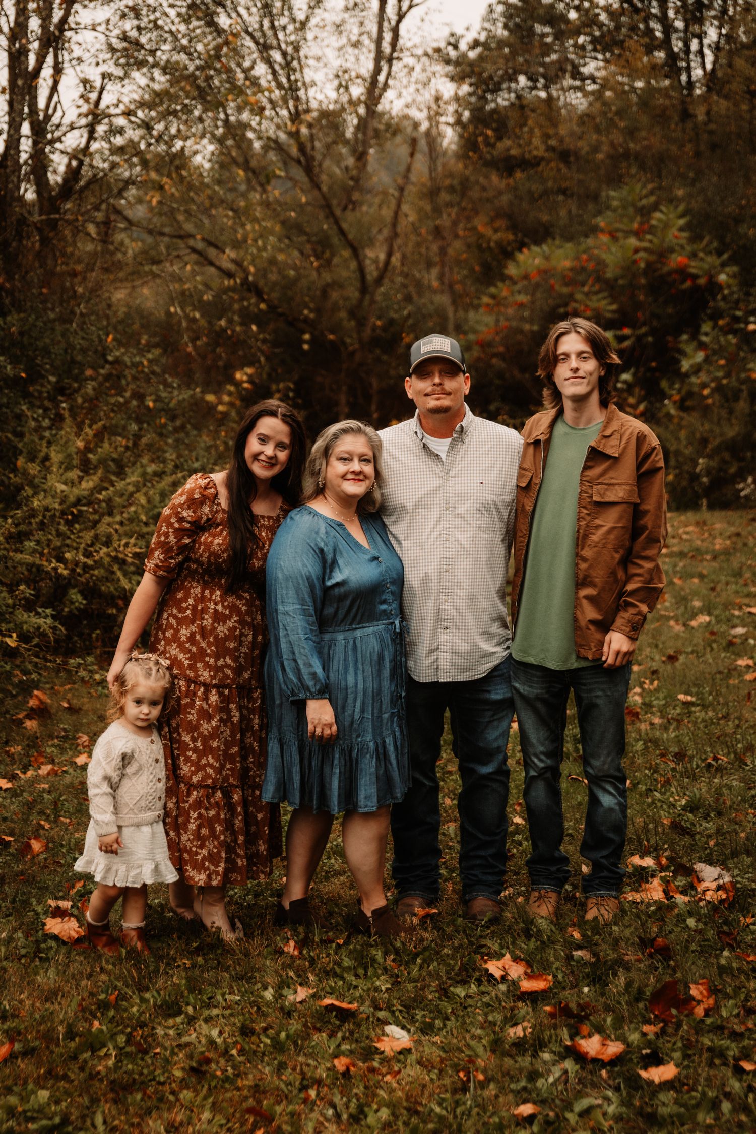A group wearing fall-colored clothing poses together outdoors among autumn trees and fallen leaves.