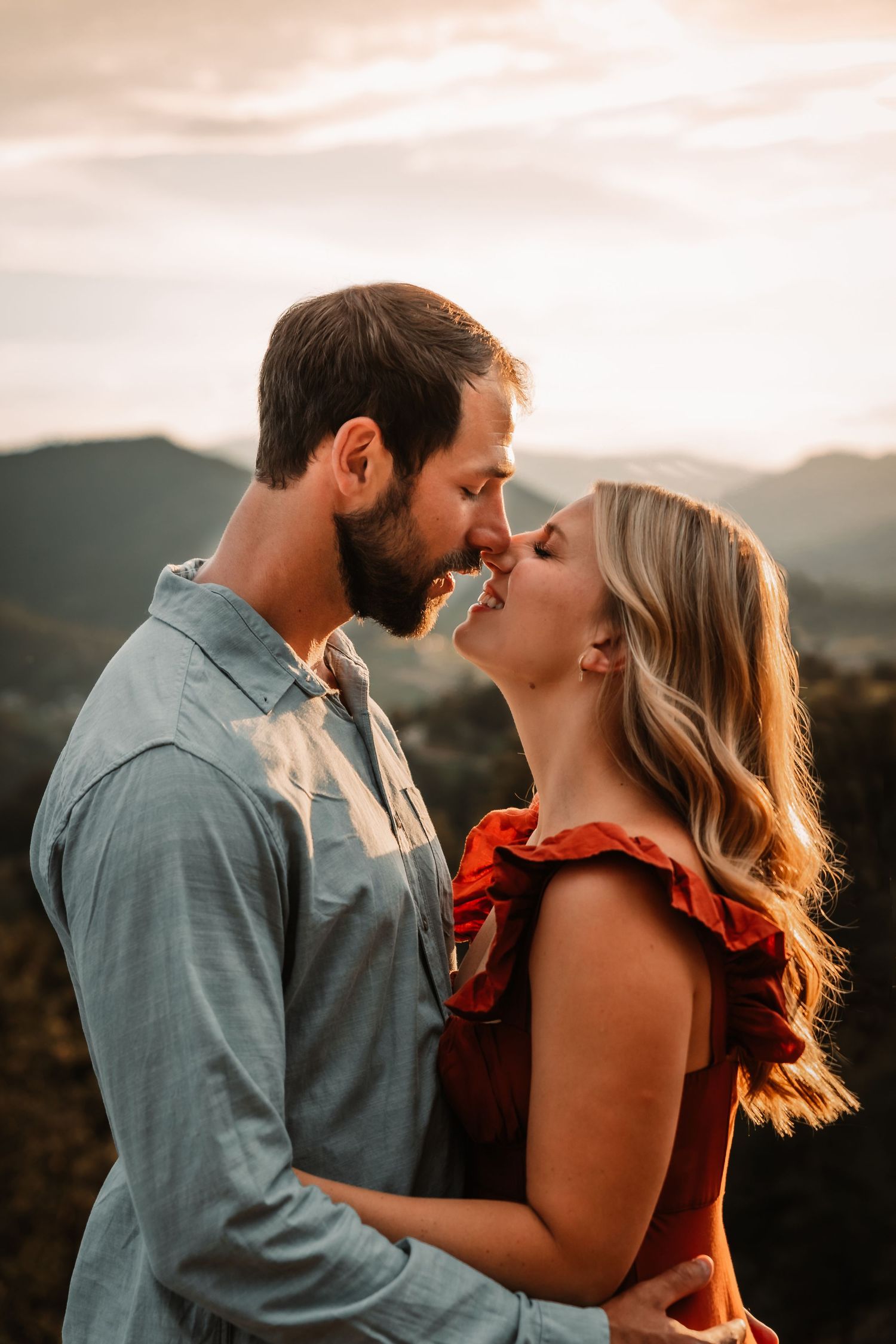 A romantic couple embraces during golden hour with mountains in the background at sunset.