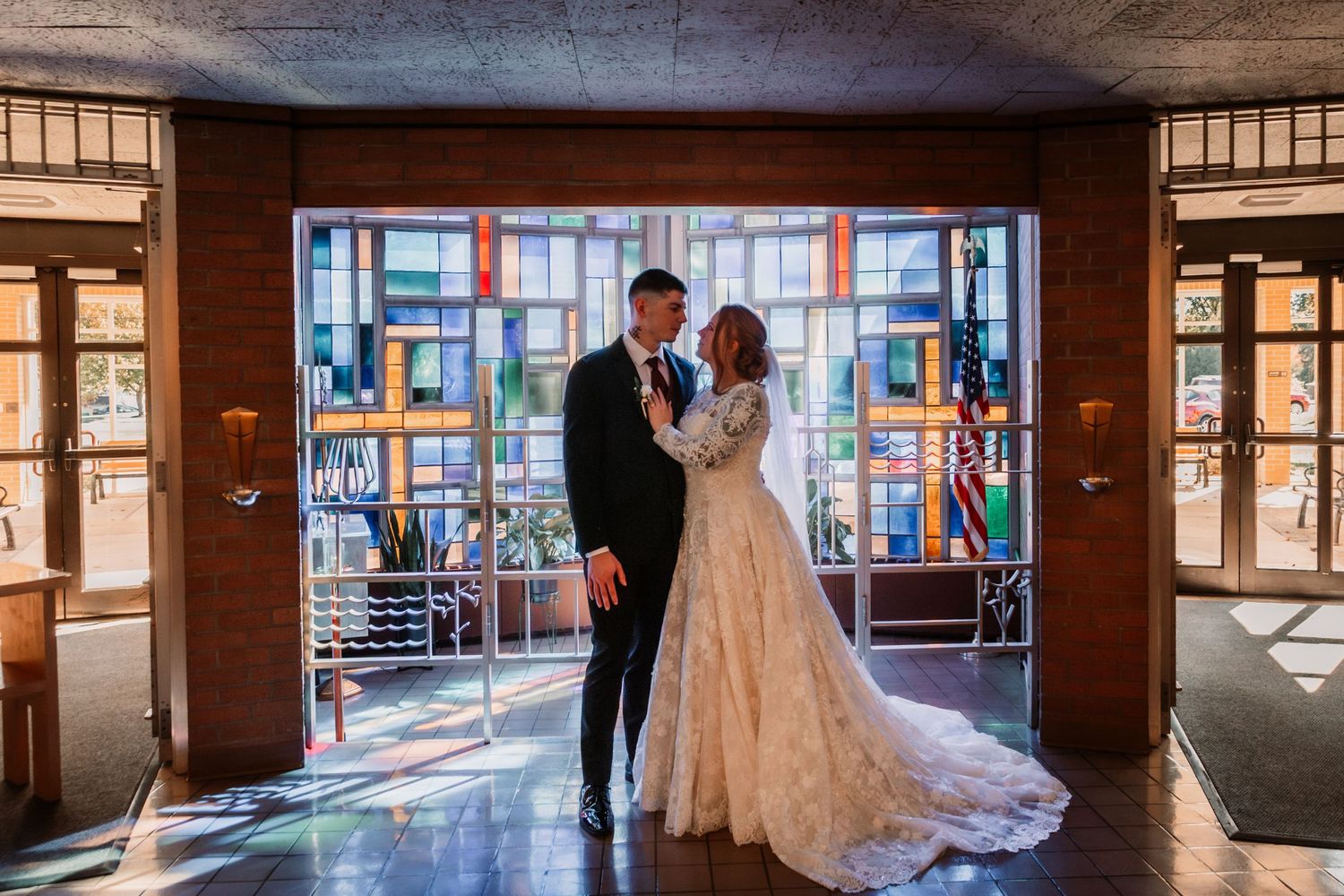 A couple in wedding attire embraces in a dark hallway with colorful stained glass windows casting vibrant light.
