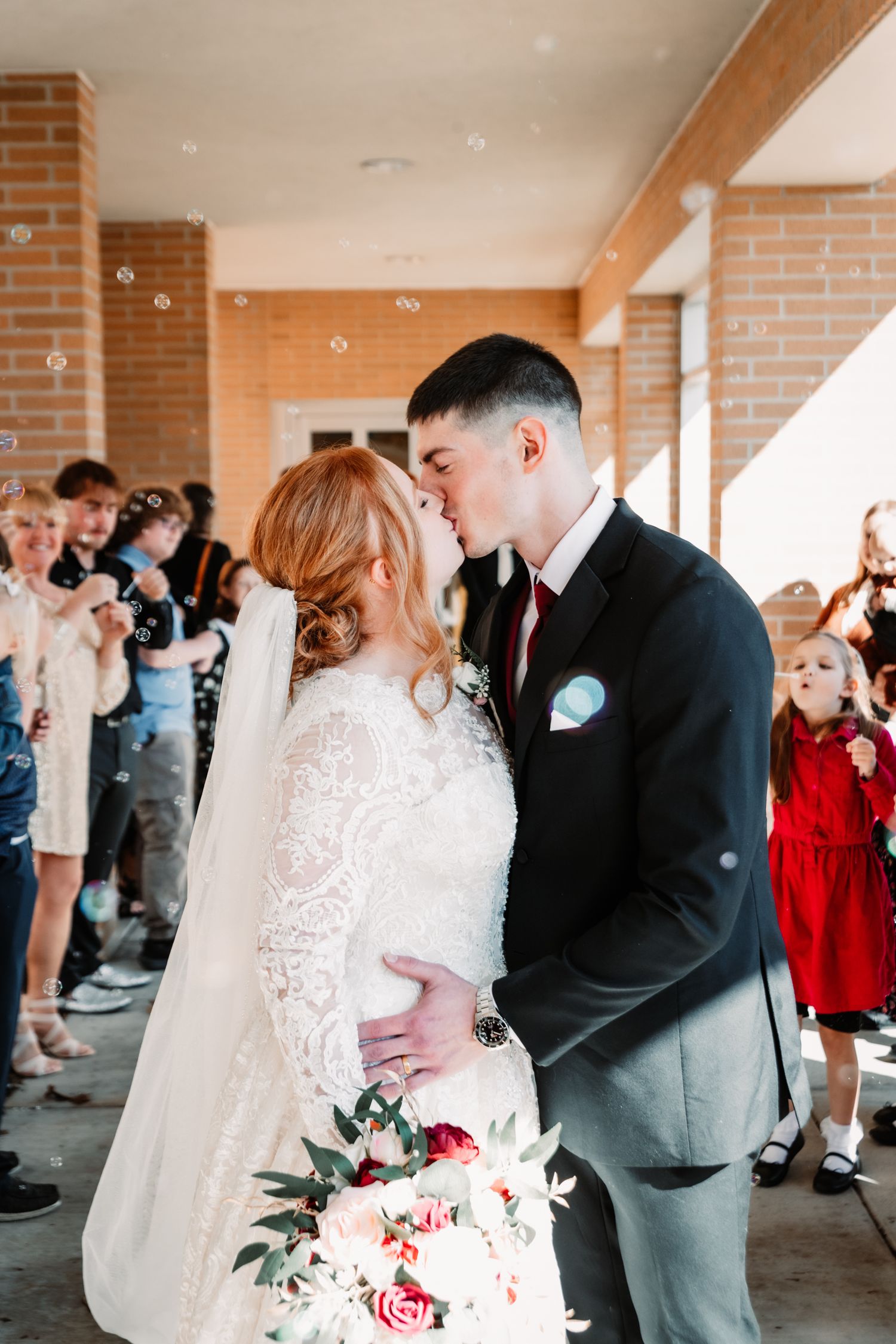 Newlywed couple shares a romantic kiss under covered walkway surrounded by guests while holding a pink and white bouquet.