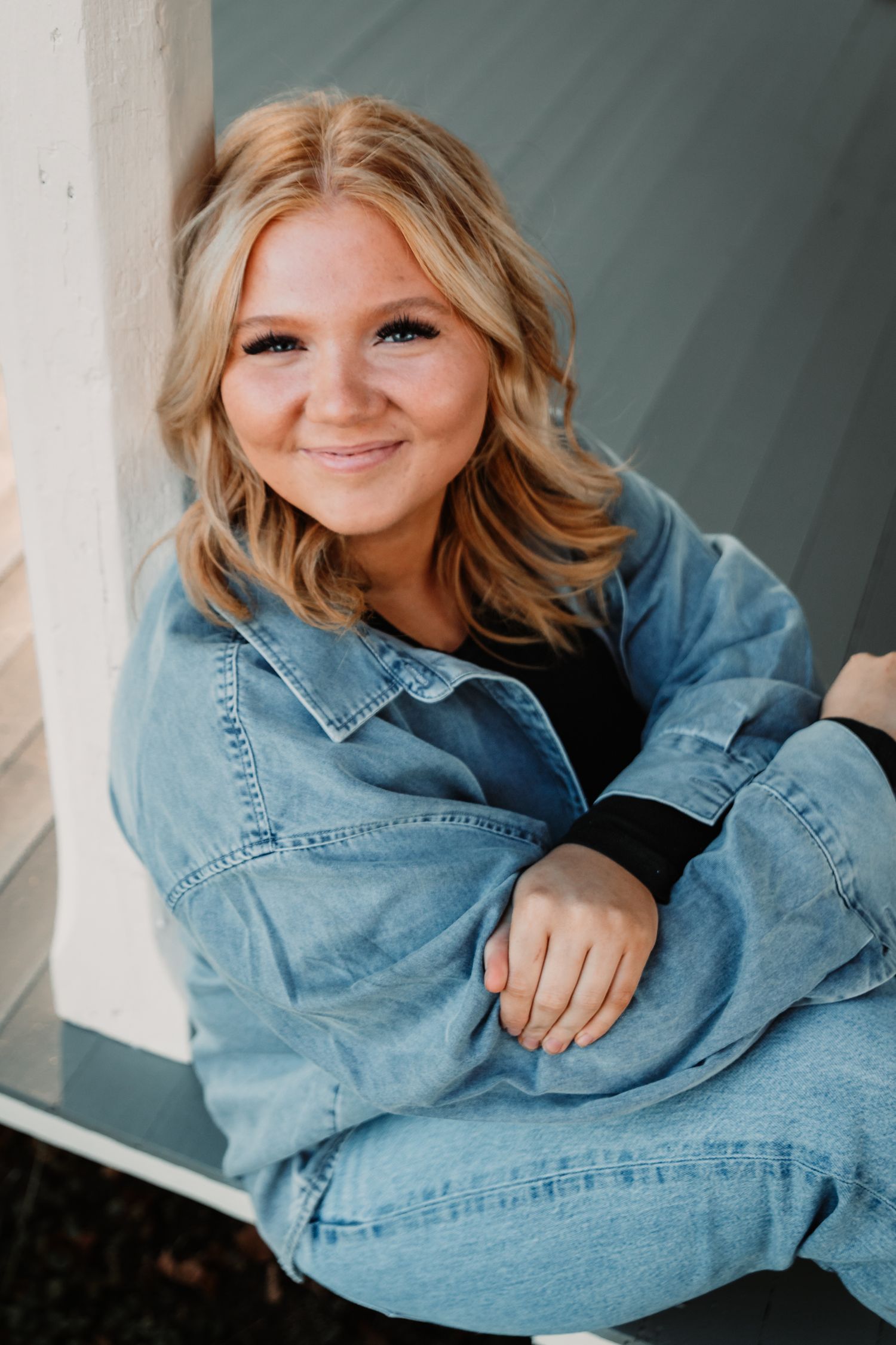 A smiling individual wearing a denim outfit sits casually against a wall in a relaxed outdoor portrait.