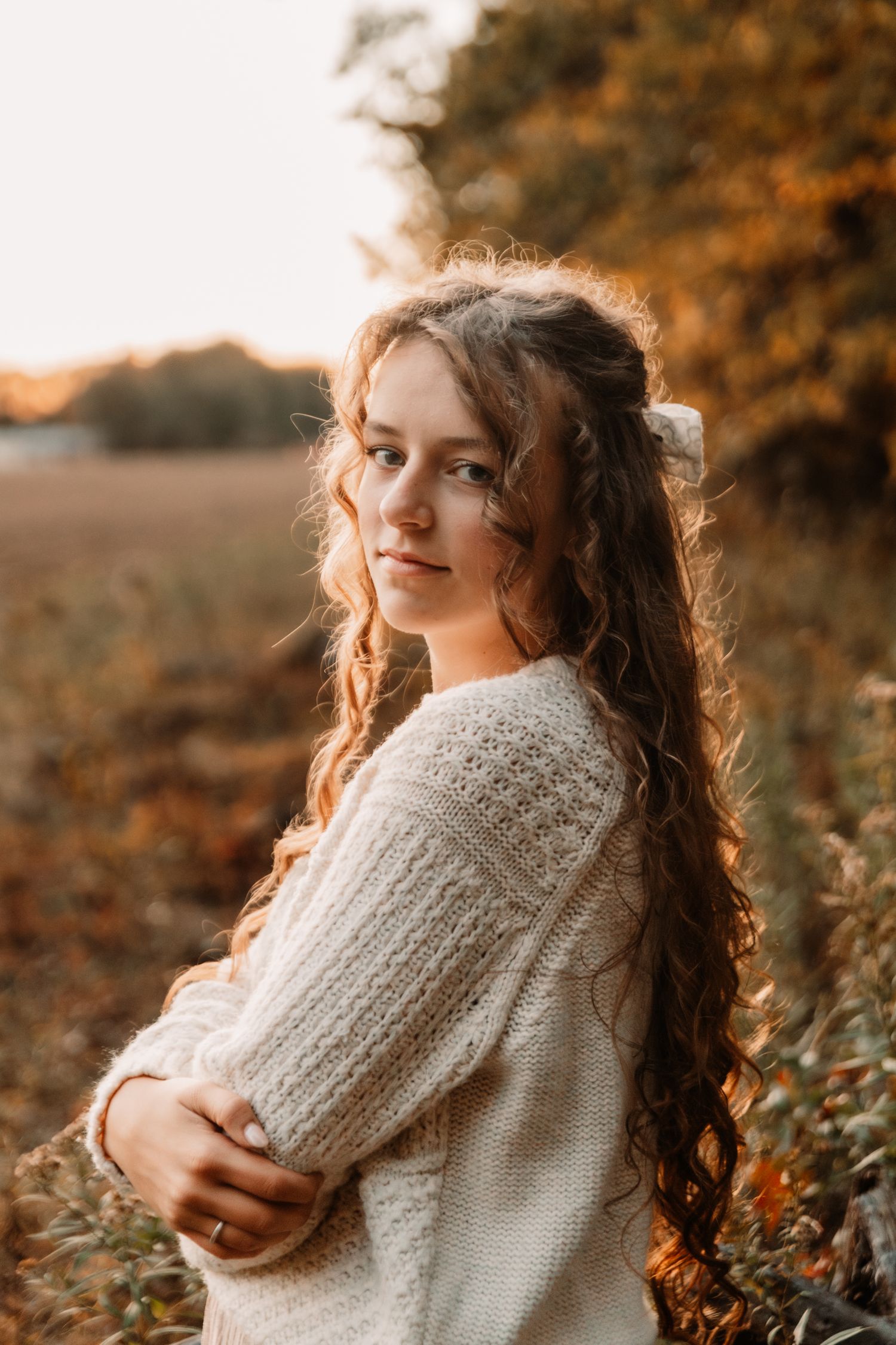 A figure in white knit sweater with flowing dark wavy hair poses at sunset in an autumn outdoor setting.