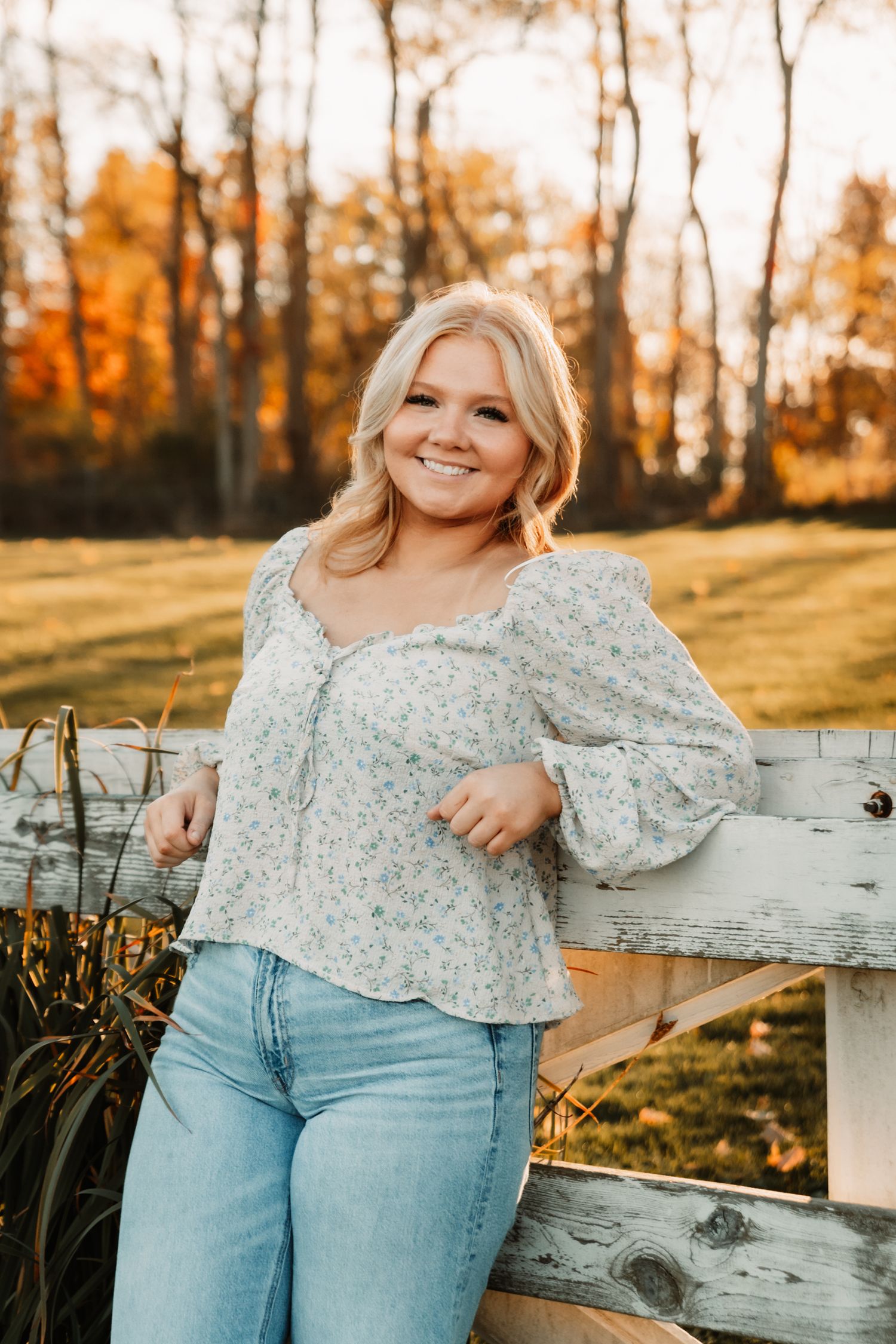 Woman in floral top and jeans poses by rustic fence during golden autumn sunset.